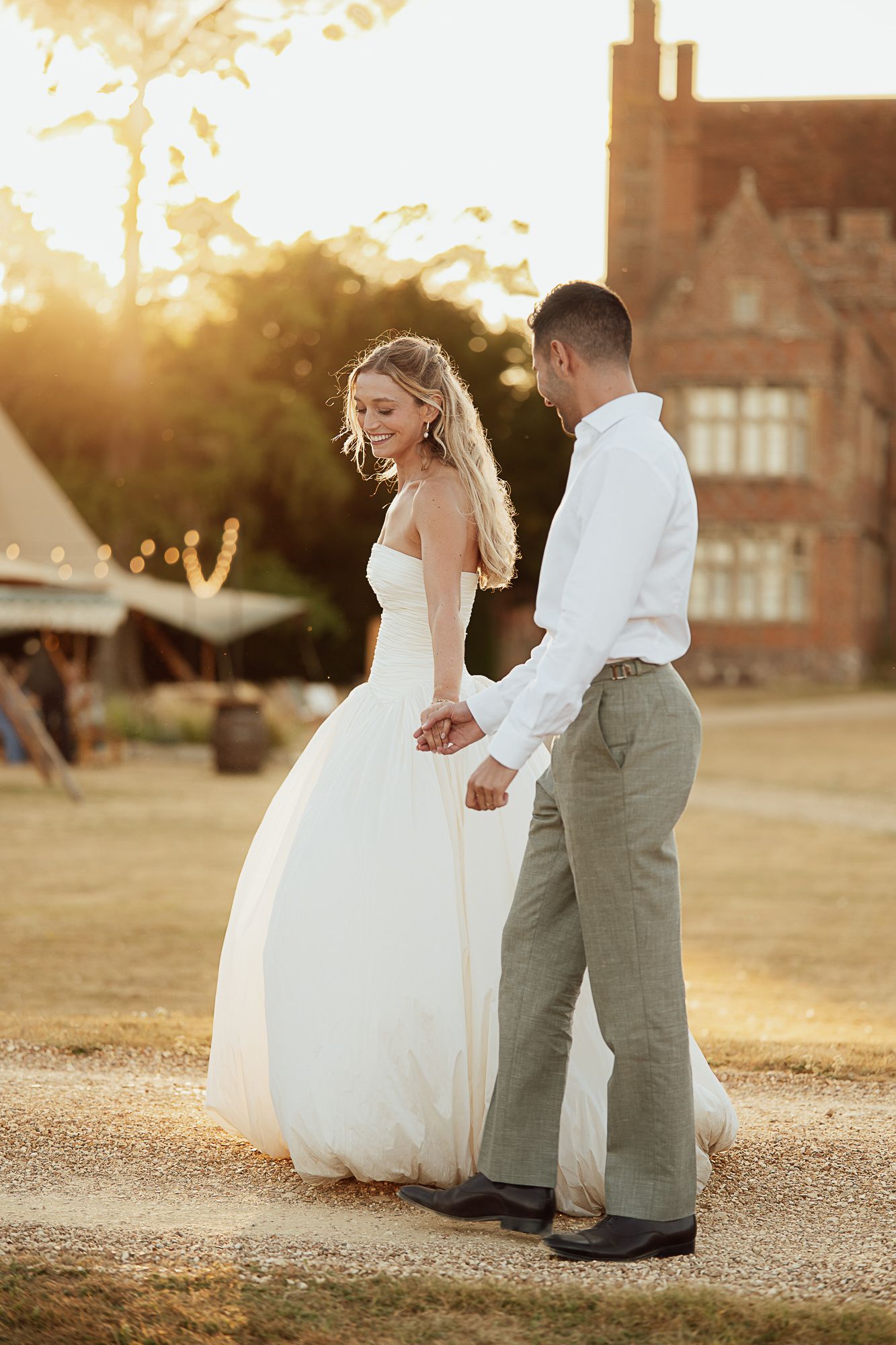 A bride and groom hold hands at golden hour, walking near a historic manor with warm sunset light glowing behind them.