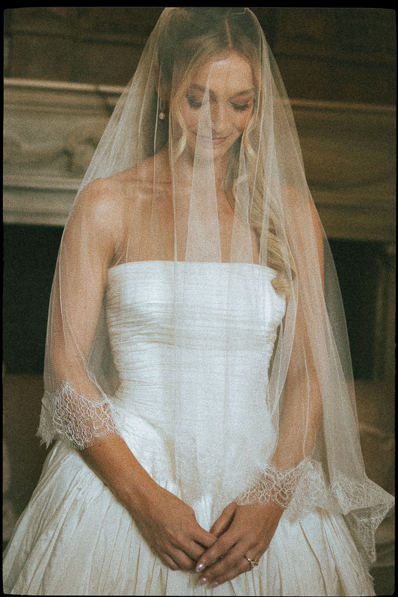 Bride in strapless white gown wears lace-trimmed veil looking down in elegant wedding portrait.