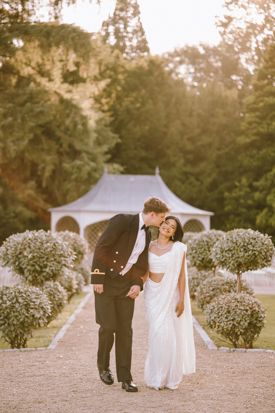 Groom in military tuxedo kisses brides cheek at golden hour in manicured garden.