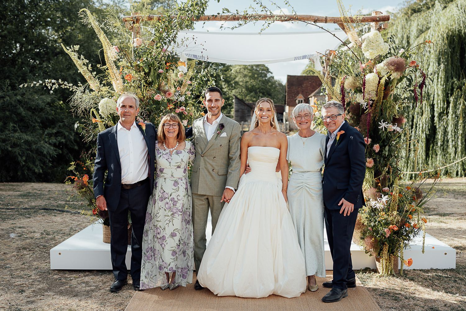 Bride and groom pose with four family members before floral arch backdrop at outdoor wedding ceremony.