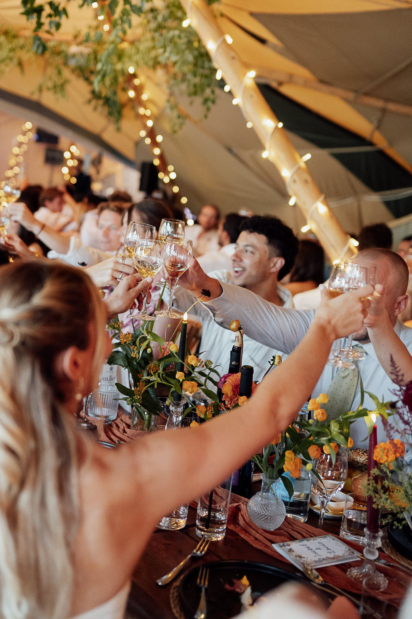 Wedding guests raising champagne glasses in a toast at a beautifully decorated outdoor reception under a tent.