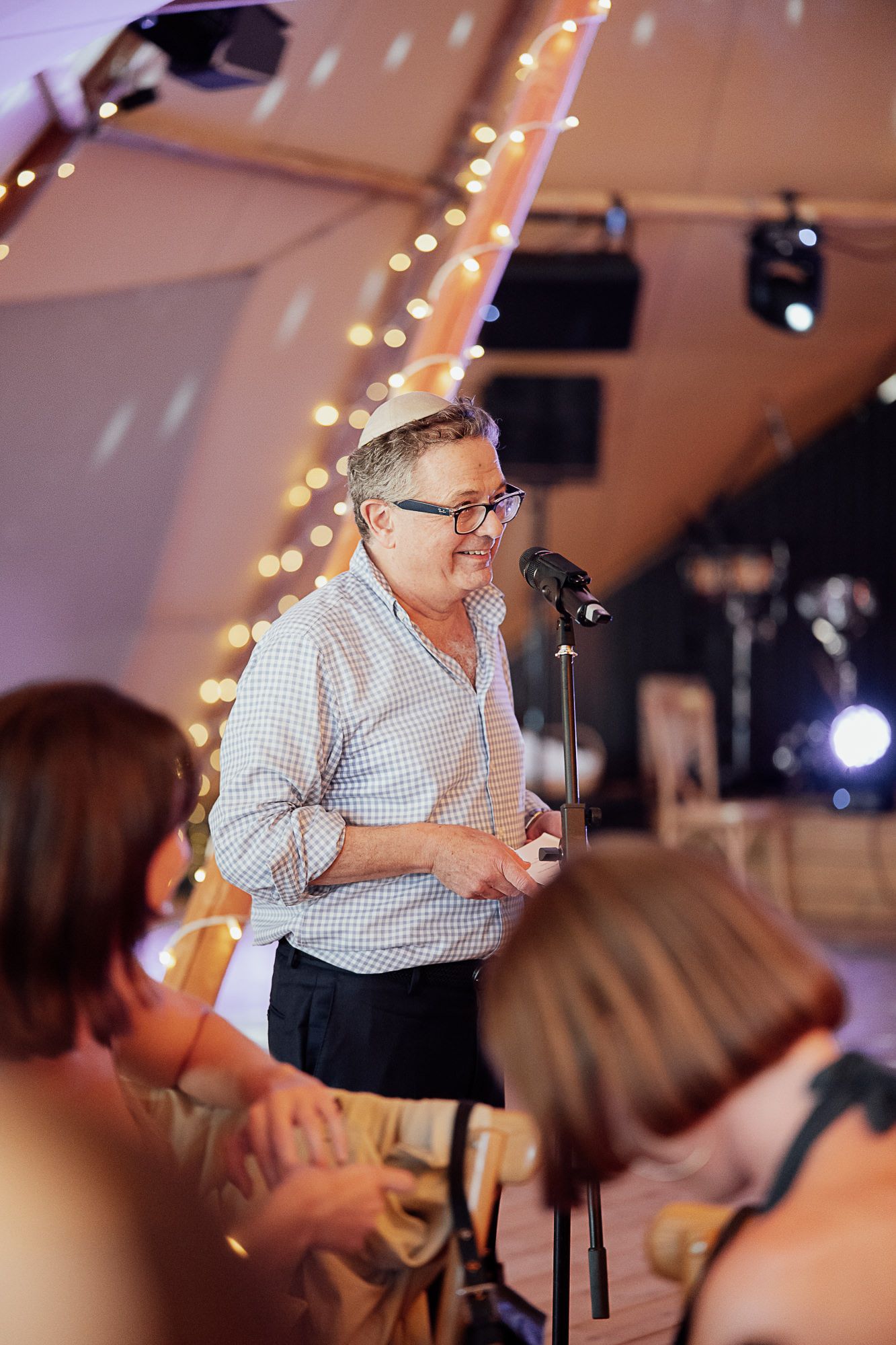Man in glasses speaking at microphone during elegant event in decorated tent with fairy lights.