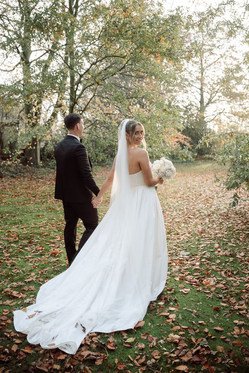A couple in wedding attire walks through autumn leaves in a wooded setting as the bride's white gown trails behind them.