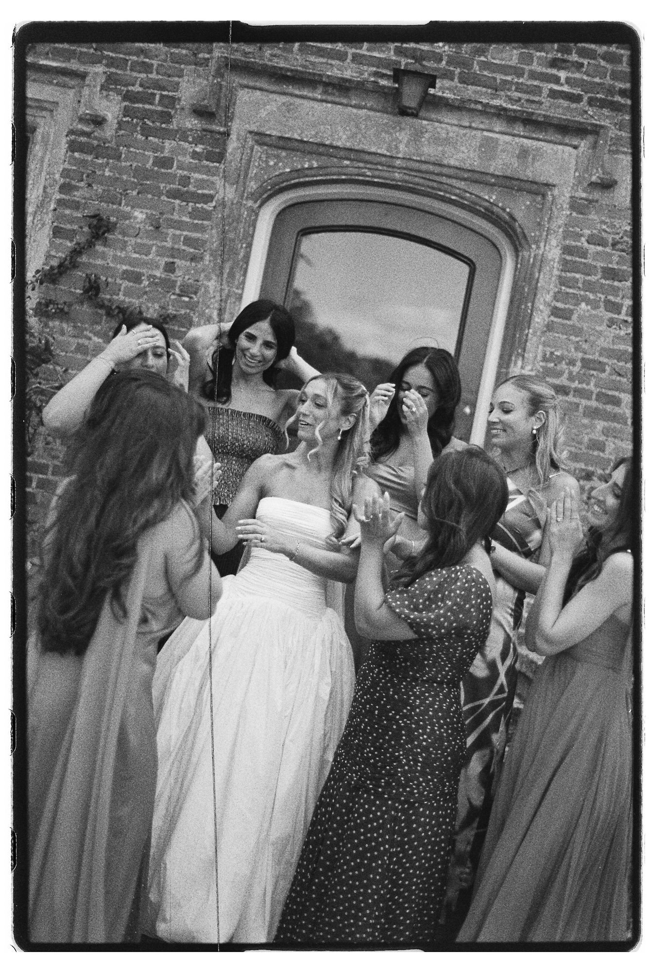 Black and white photo of a bride in white strapless gown surrounded by bridesmaids laughing outside a brick building.