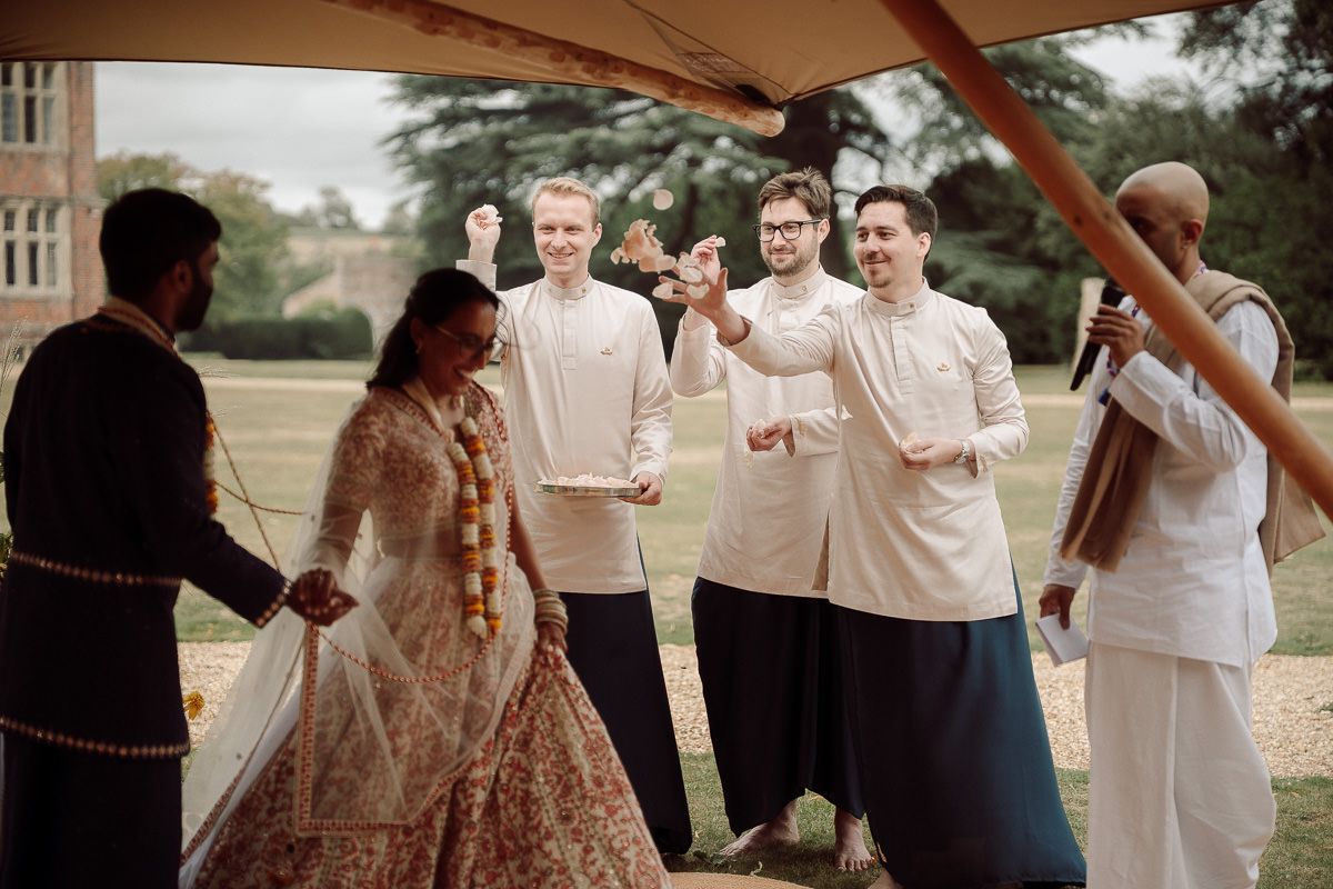 Wedding guests gather outdoors on a sunny day, holding papers and mingling near a brick building.