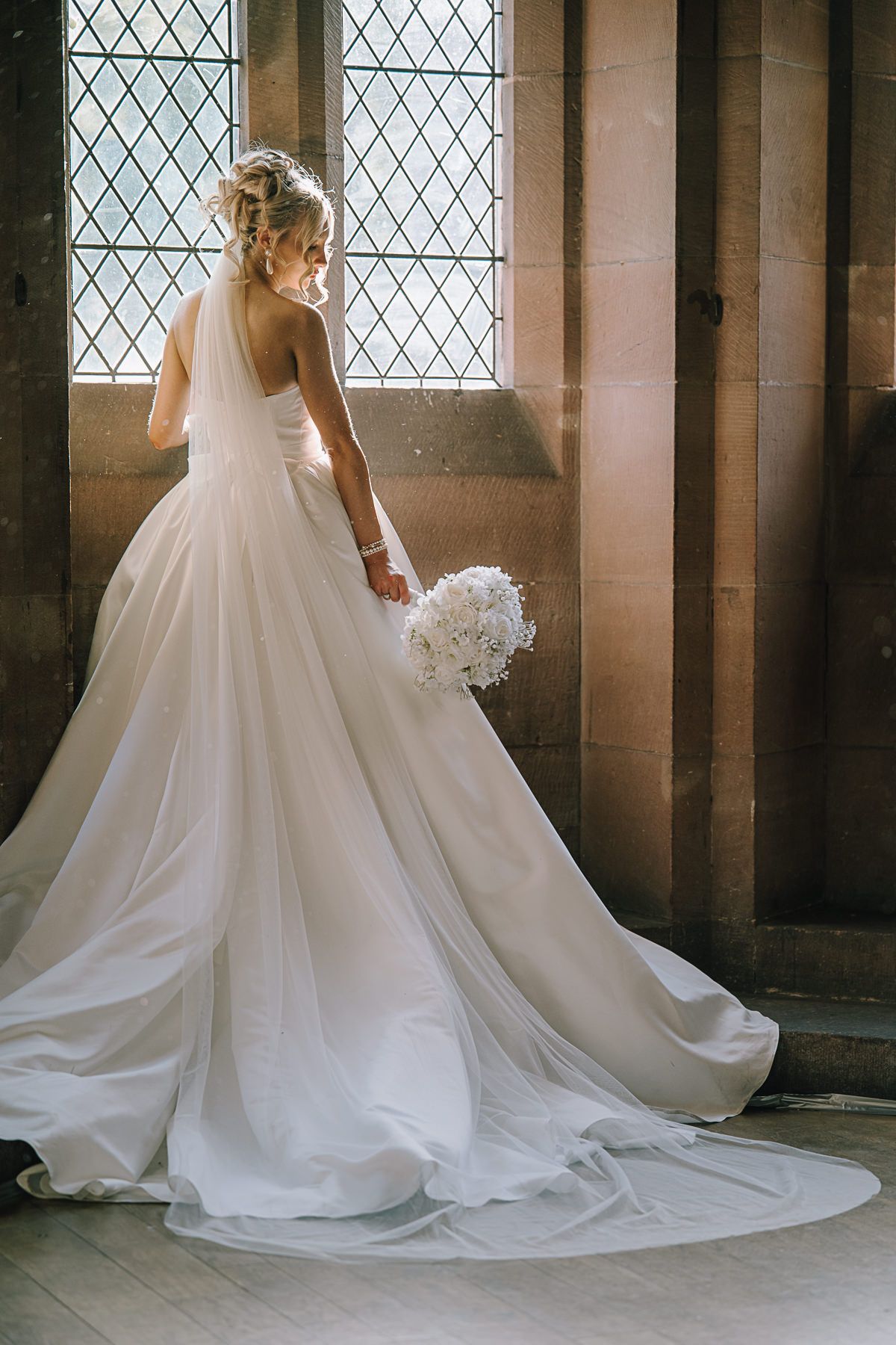 Bride in elegant white ball gown with long train holds bouquet, standing by leaded glass window in stone venue.