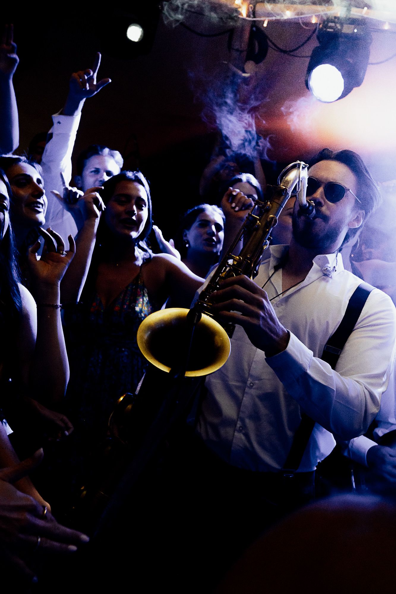 Saxophonist in white shirt and sunglasses performs for an energetic crowd at a smoky nightclub event.