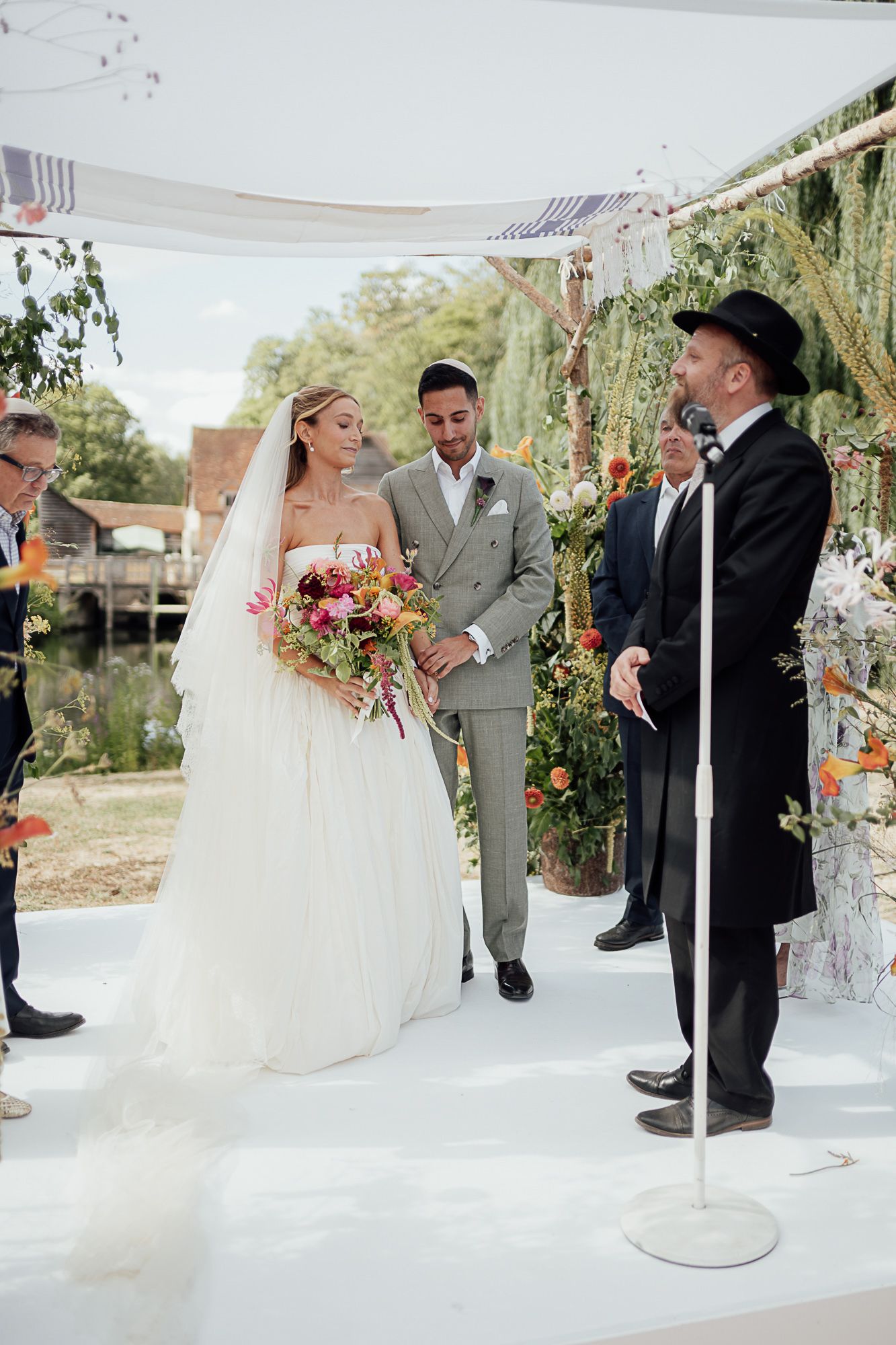 Bride and groom exchange vows outdoors under a floral chuppah, officiated by a rabbi at an elegant garden wedding.