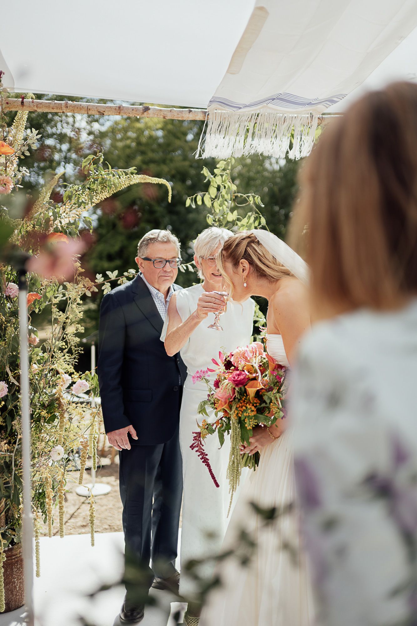 Bride holding colorful bouquet stands with groom under floral wedding arch during outdoor ceremony.