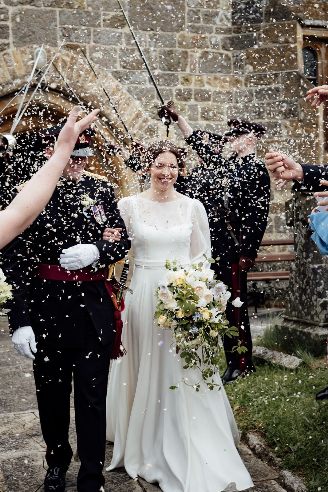 A couple walks through falling confetti after their wedding ceremony with guests celebrating beside a stone church wall.