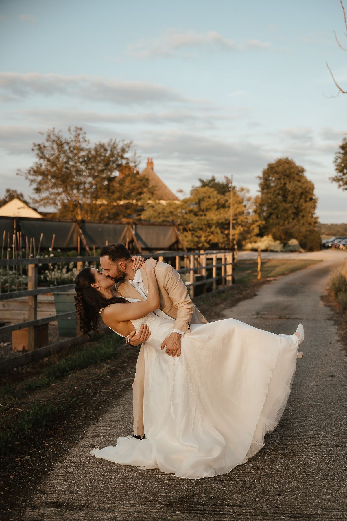 A romantic couple in wedding attire share intimate moments at sunset on a rustic country road near rural buildings.