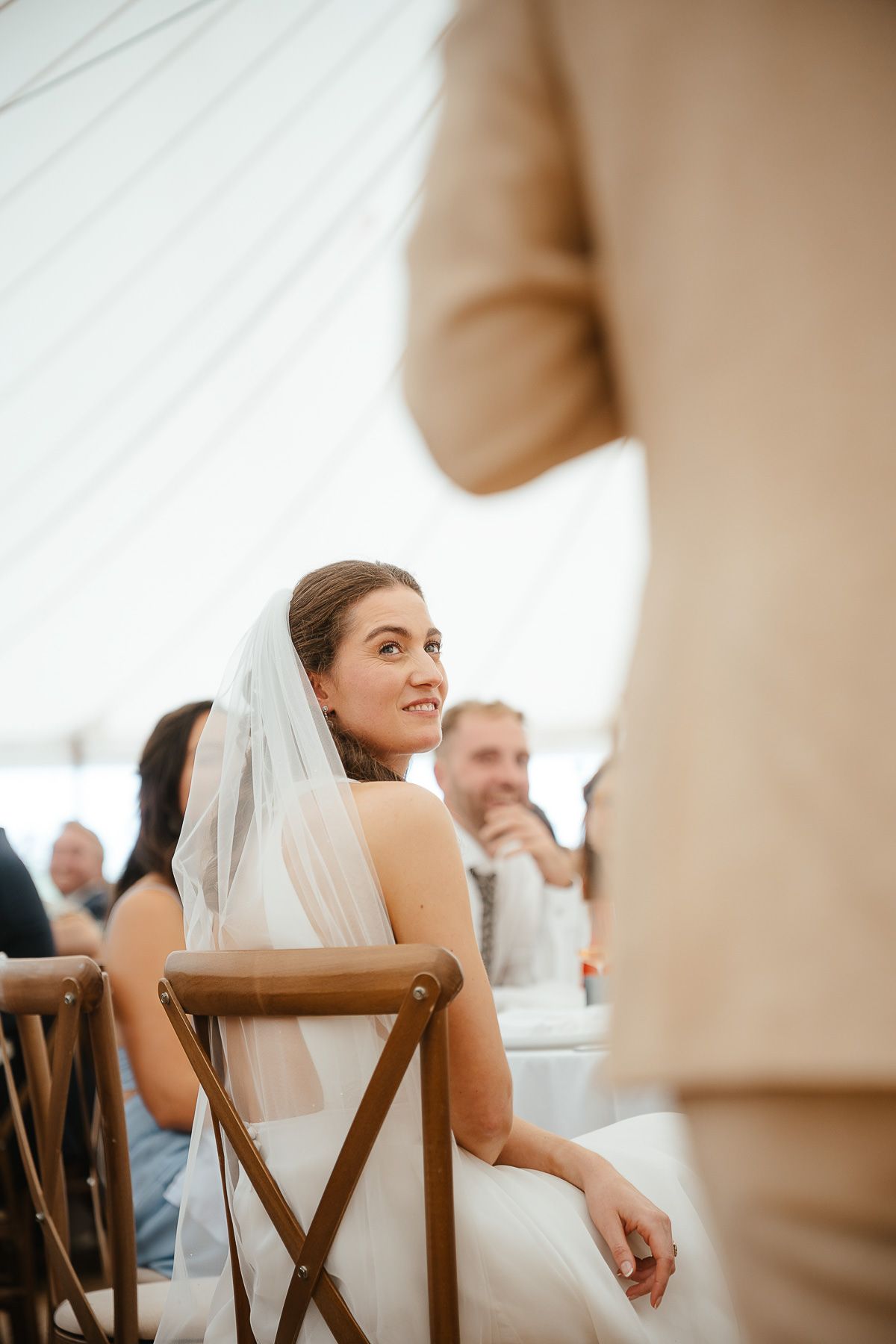 A bride in a white wedding veil smiles during an indoor reception while seated at a wooden dining chair.