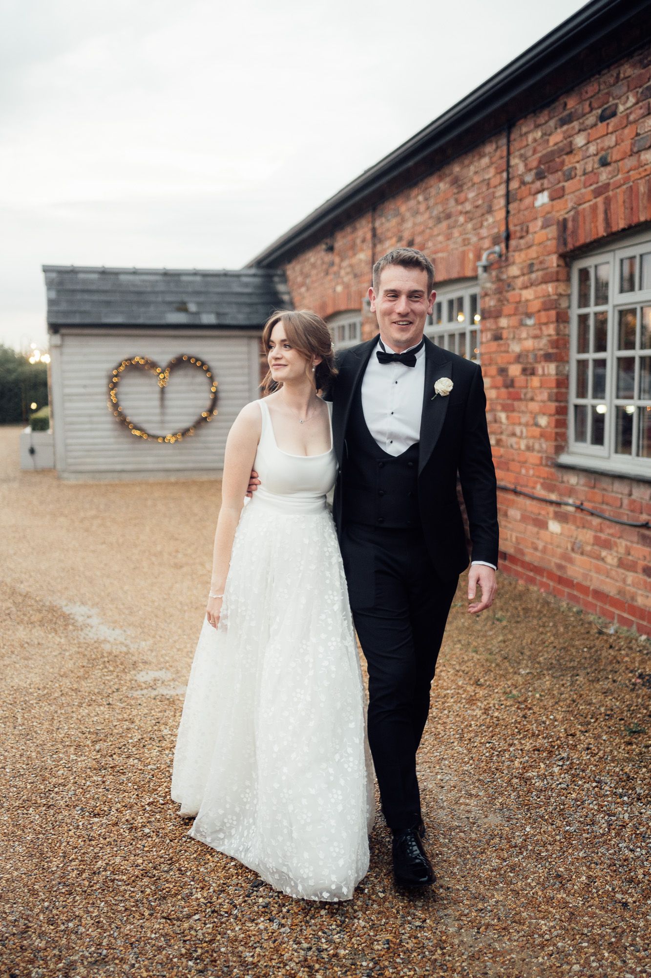 Happy couple in wedding attire walking and laughing together outside a brick building on a gravel path.