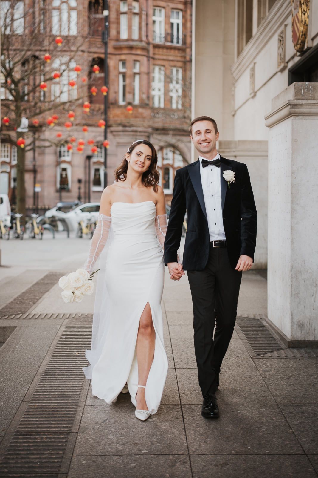 A bride in a white slit dress and groom in classic black tuxedo hold hands while walking on a city sidewalk.