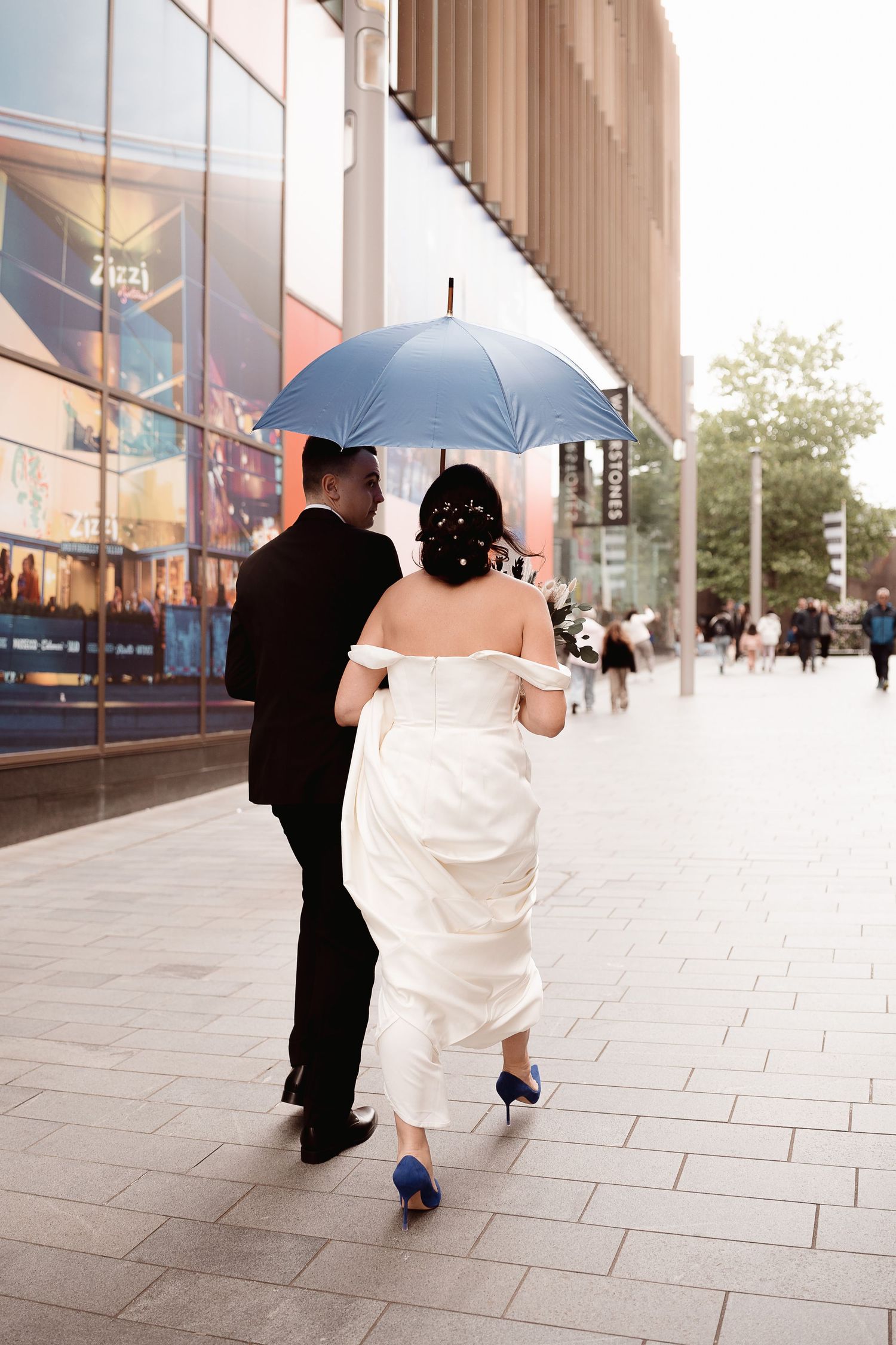 A couple walking on an urban sidewalk, with the bride in a white gown and groom holding a blue umbrella.