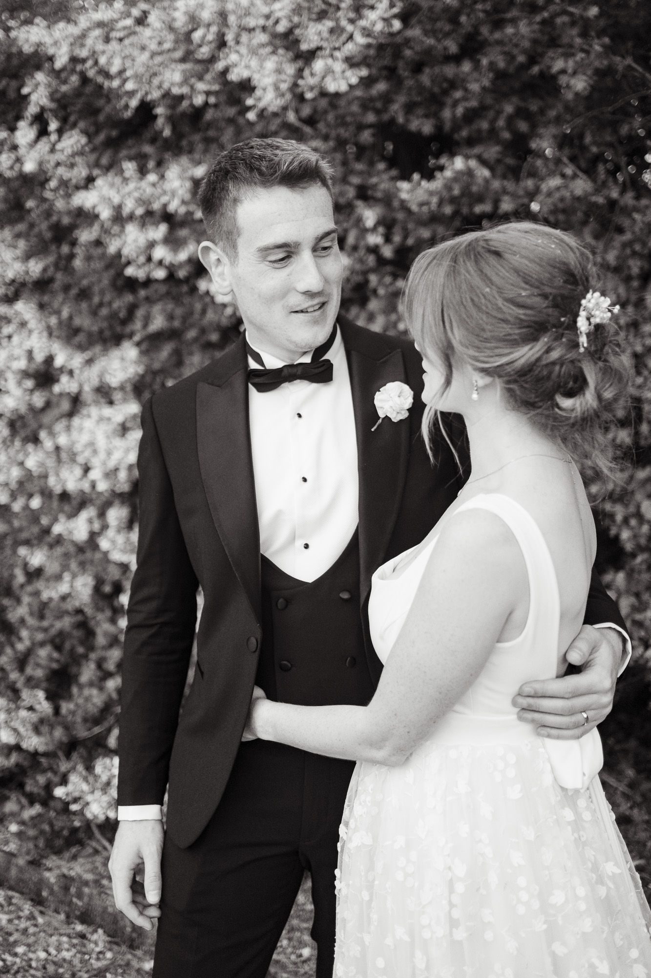 A black and white wedding photograph showing a tender moment between newlyweds in formal attire outdoors.