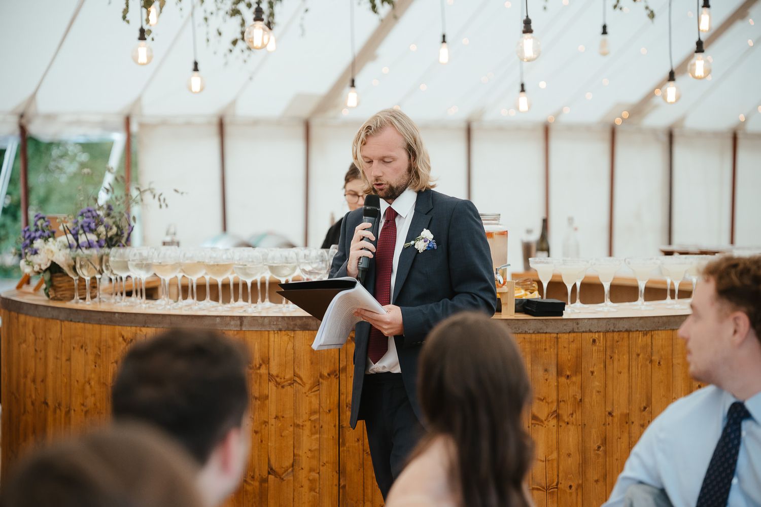 Someone in a dark suit gives a wedding speech at a reception under a white tent with wooden bar and hanging lights.