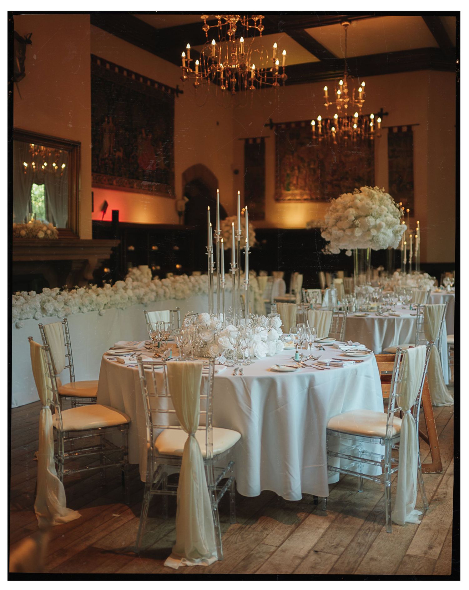 Elegant wedding reception hall with white floral centerpieces, chiavari chairs, and glowing chandeliers on hardwood floors.