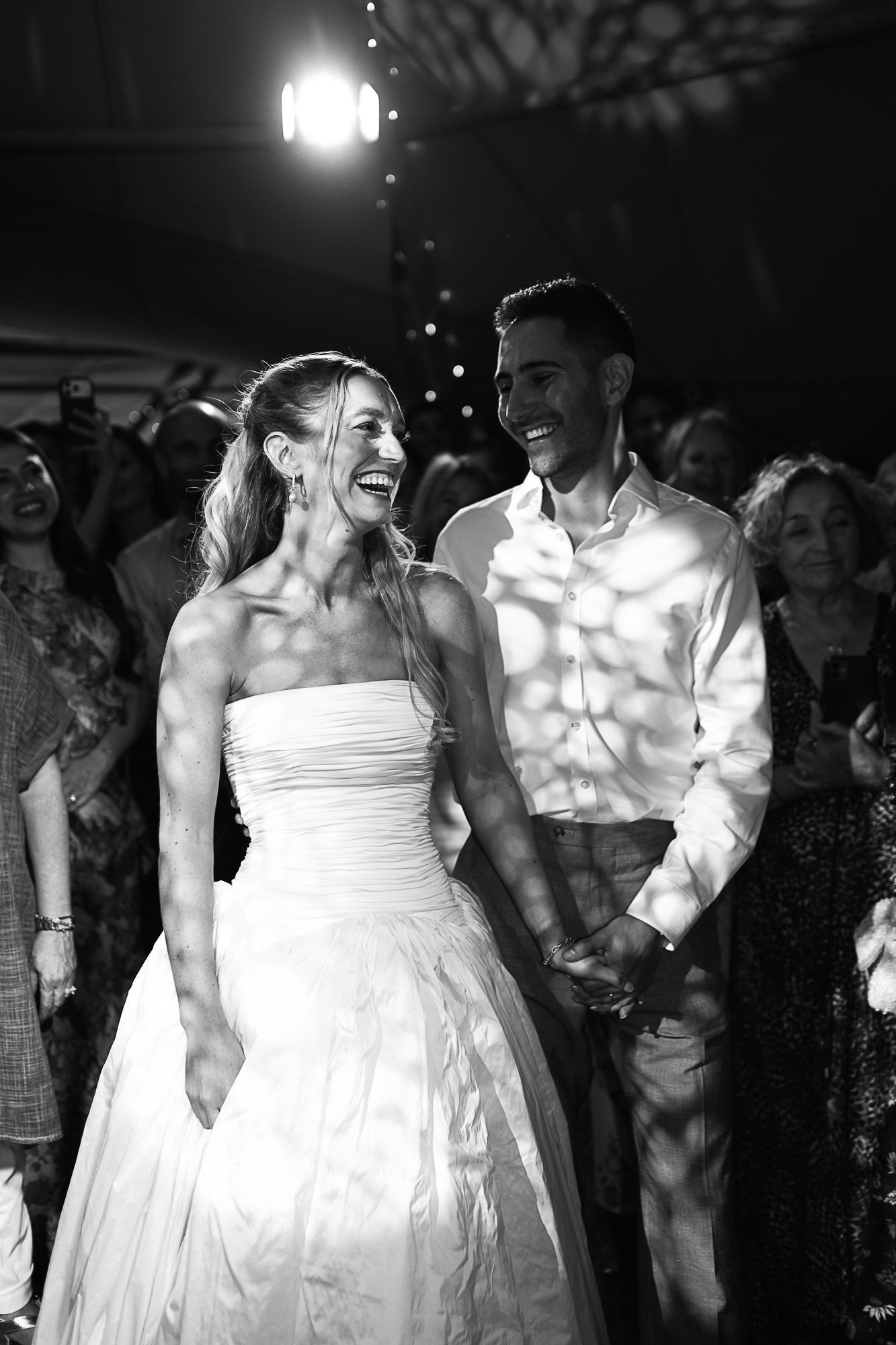 Black and white photo of a laughing bride and groom holding hands on the dance floor at their wedding reception.