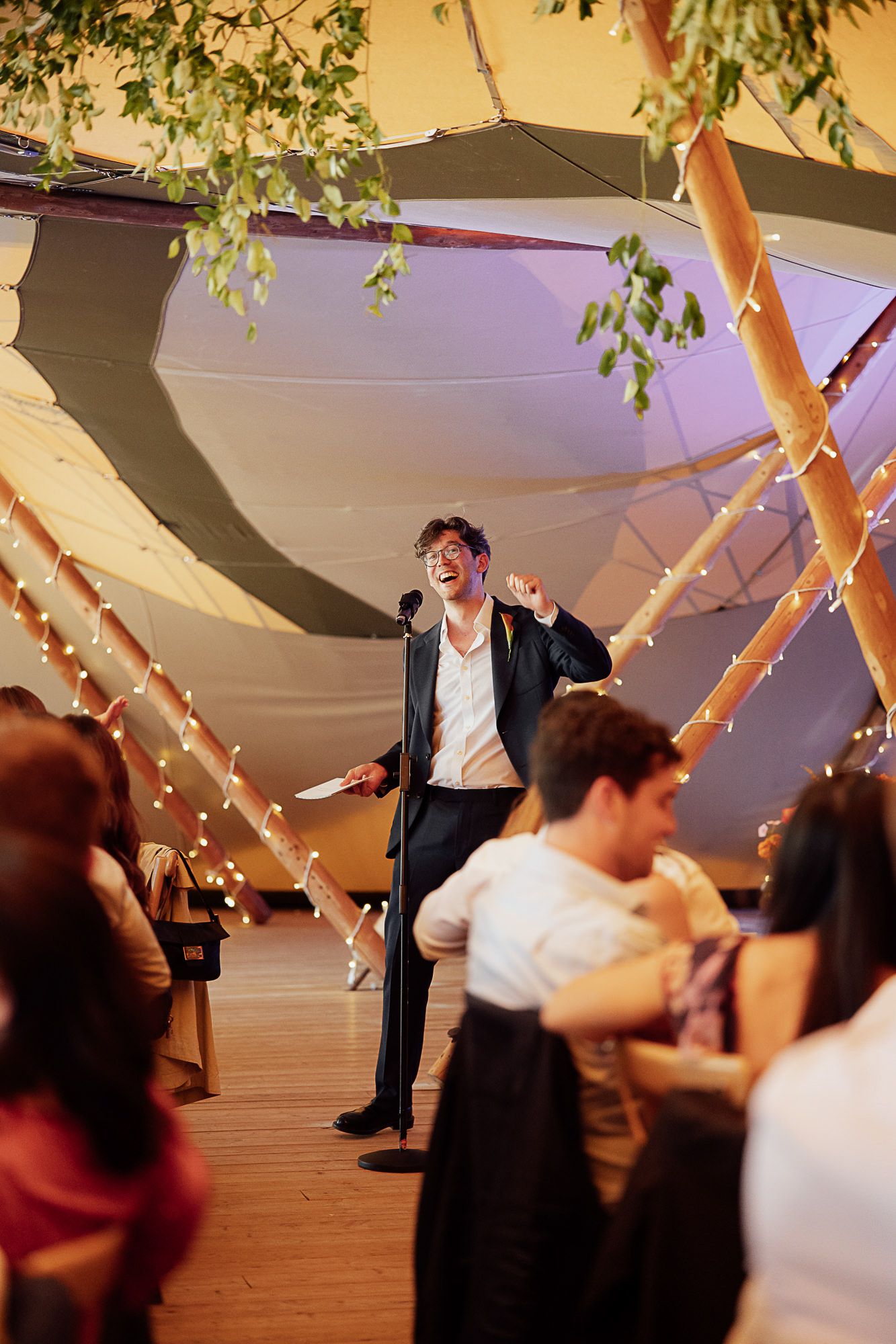 Man in suit giving an enthusiastic speech at a wedding reception inside a decorated tent with fairy lights.
