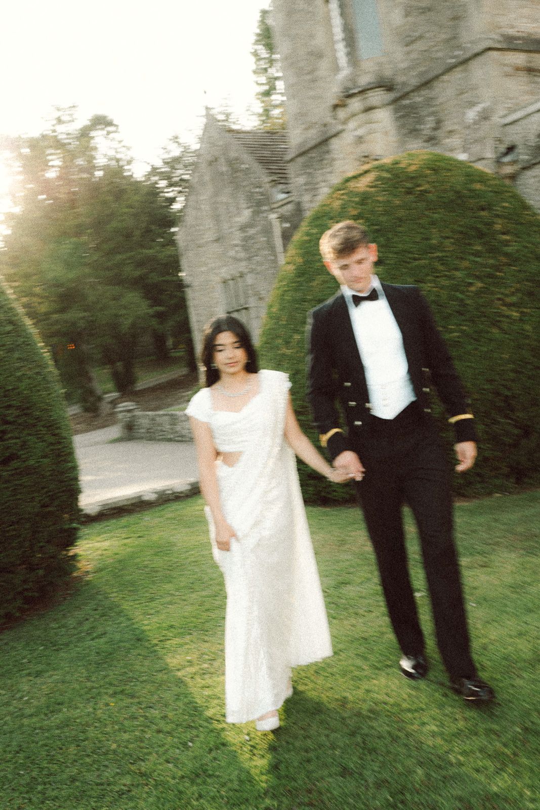 Elegant bride and groom walk hand in hand across lush green lawn beside a stone castle venue.