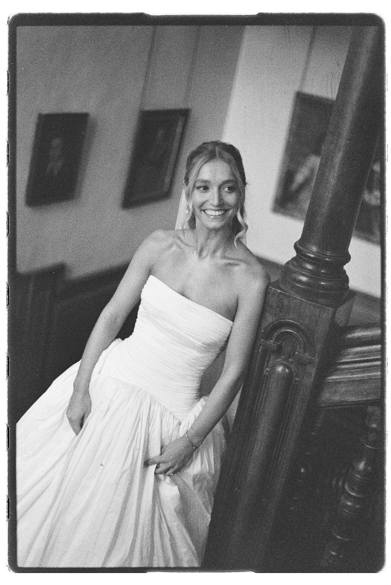 Black and white photo of a smiling bride in a strapless white gown leaning against a wooden staircase banister.
