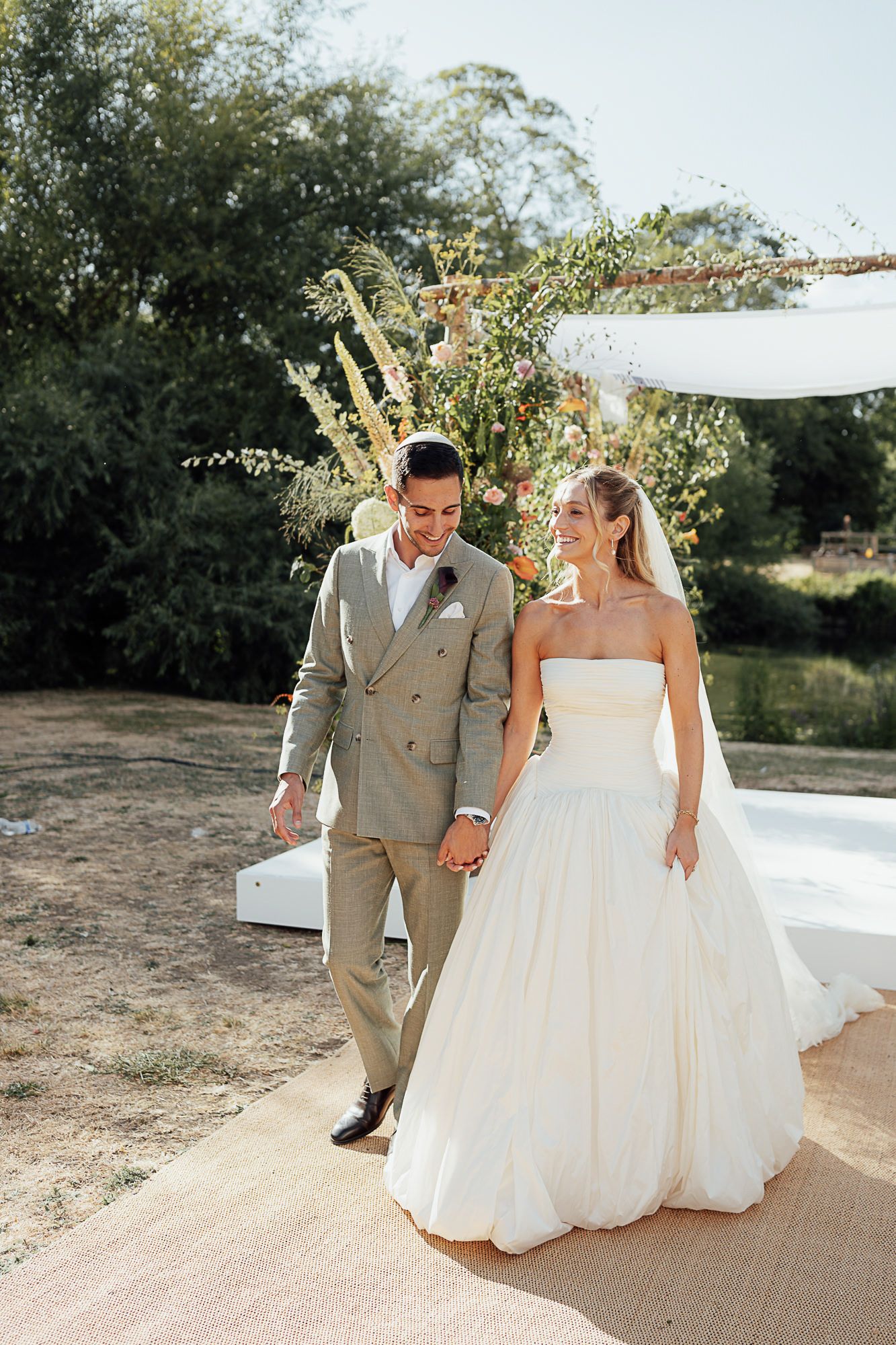 Bride in strapless white ballgown and groom in tan suit hold hands walking outdoors after wedding ceremony.