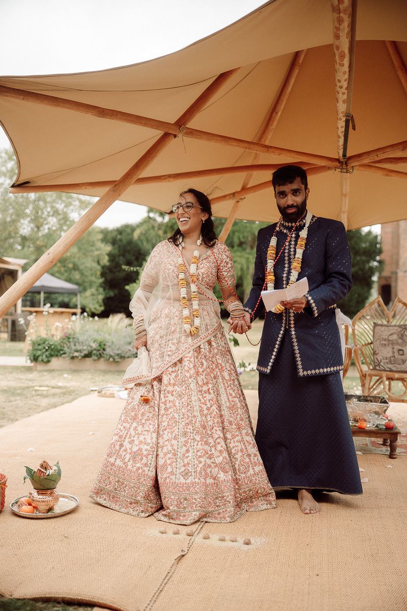 A couple in traditional South Asian wedding attire sits together under a beige tent at an outdoor celebration.