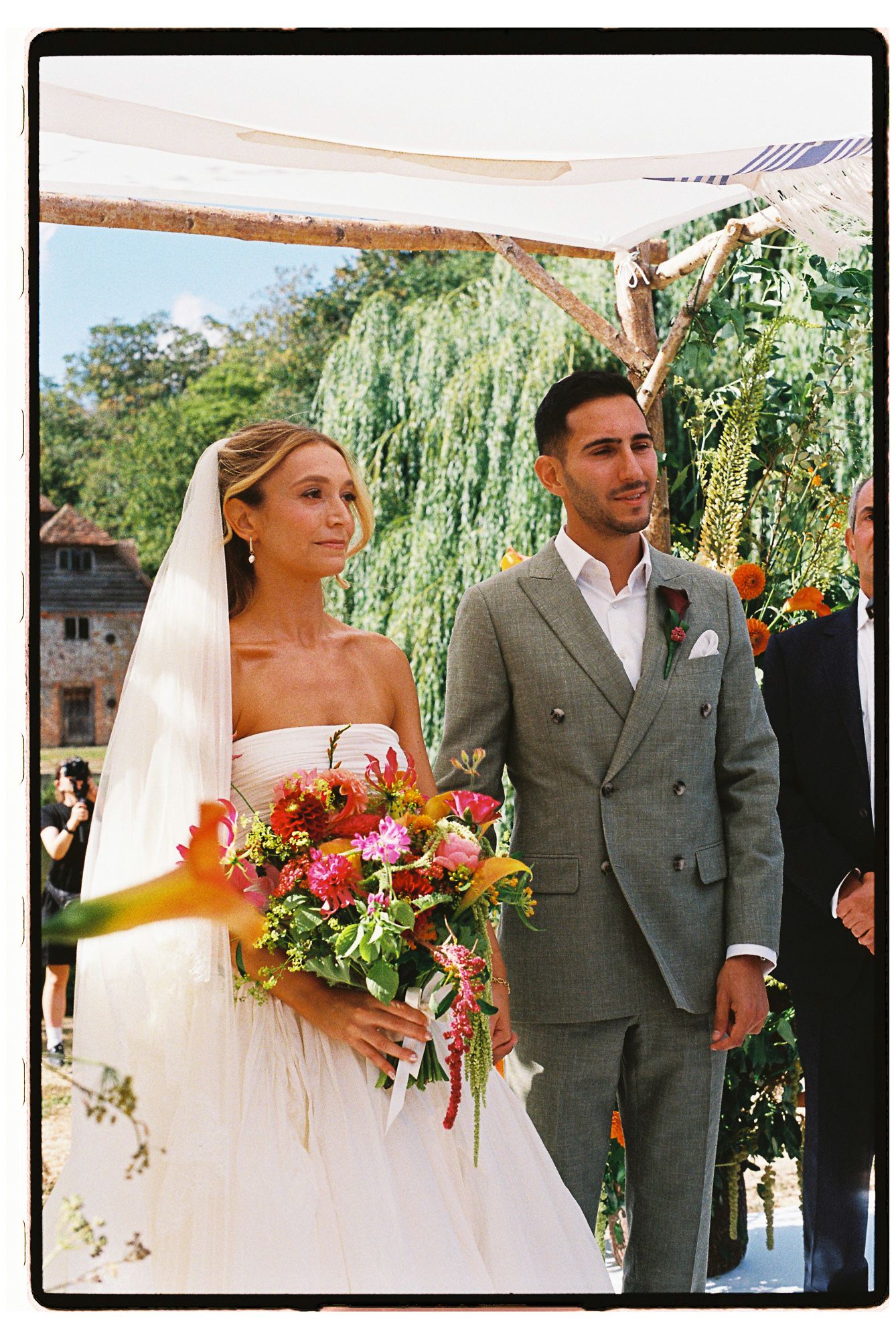 Bride in white strapless gown holding colorful bouquet stands with groom in gray suit at outdoor wedding ceremony.