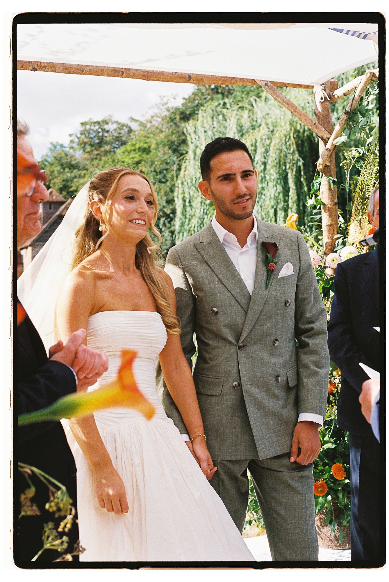Bride in white strapless gown and groom in gray double-breasted suit hold hands at outdoor wedding ceremony.