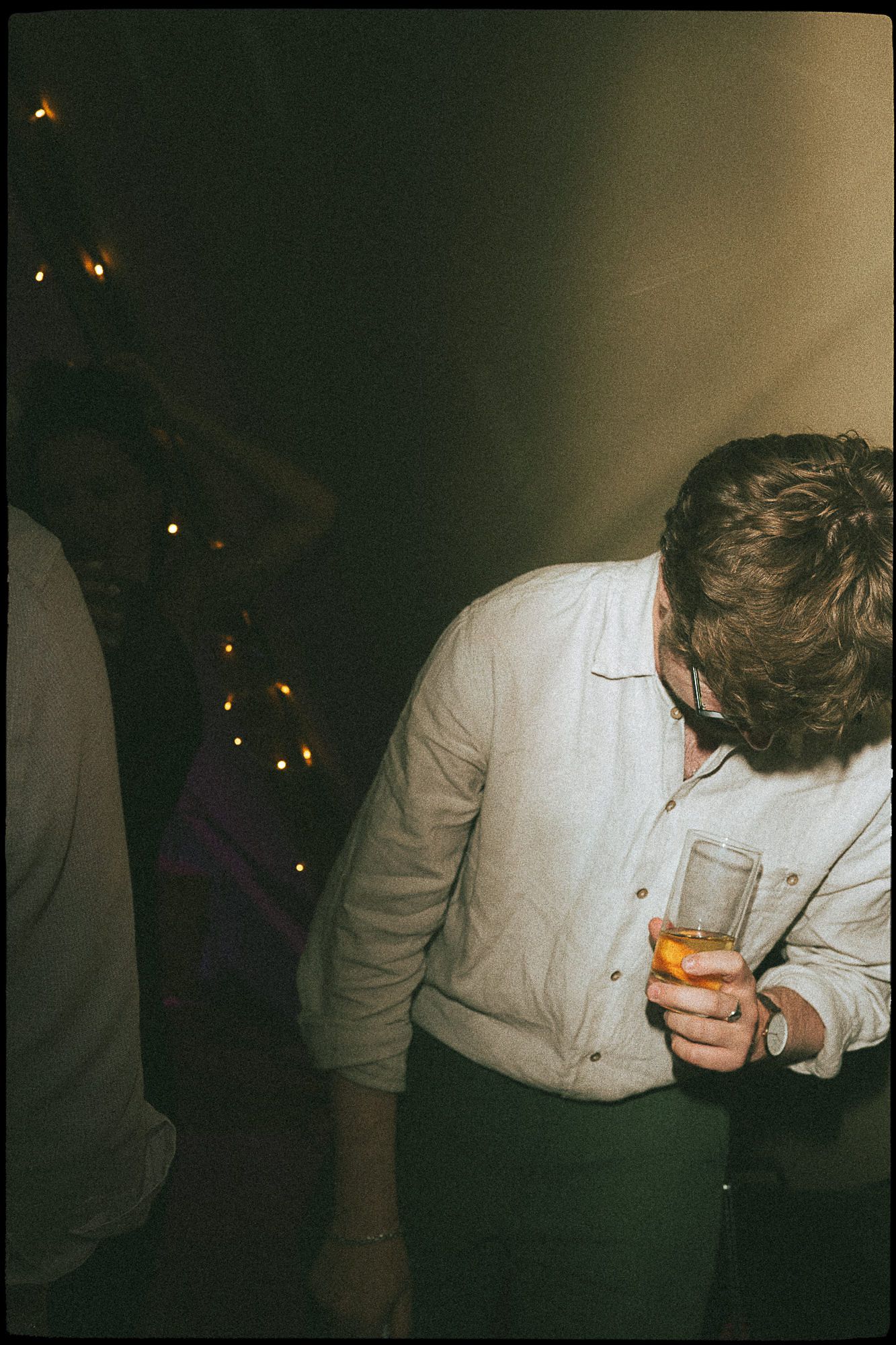 Man in white shirt holding a glass drink at a dimly lit party with string lights in the background.