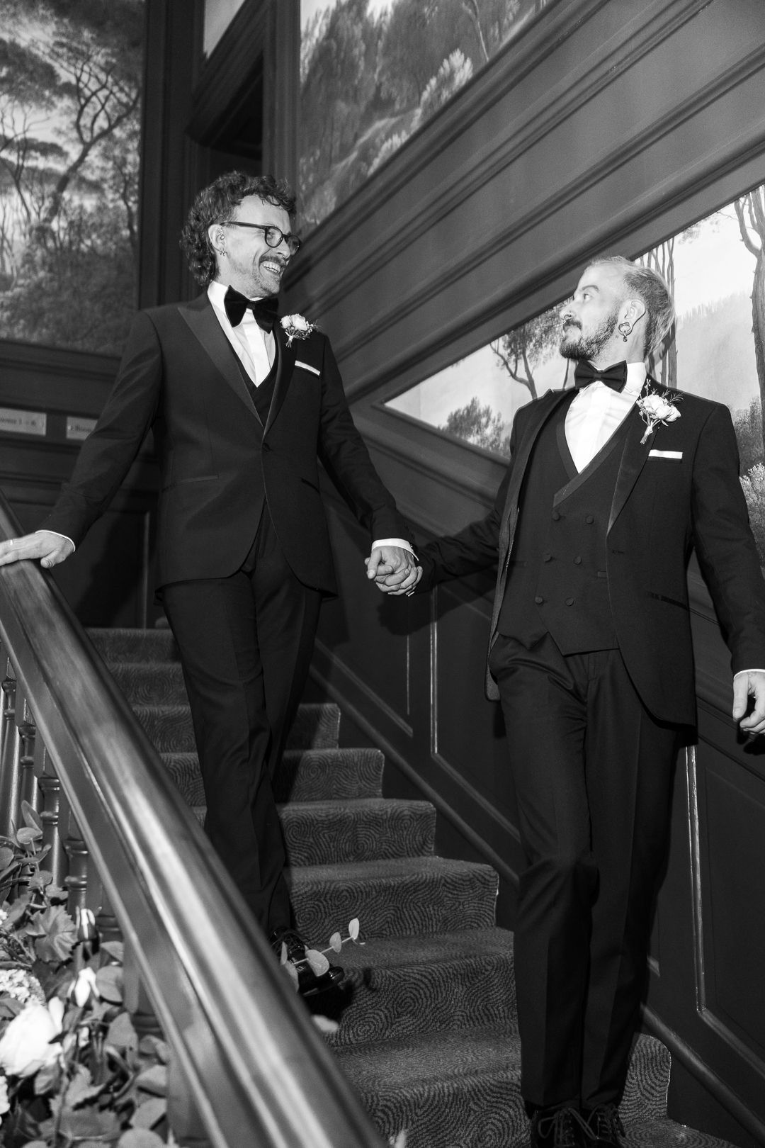 Two people in formal black tuxedos hold hands while standing on elegant staircase in black and white photo.