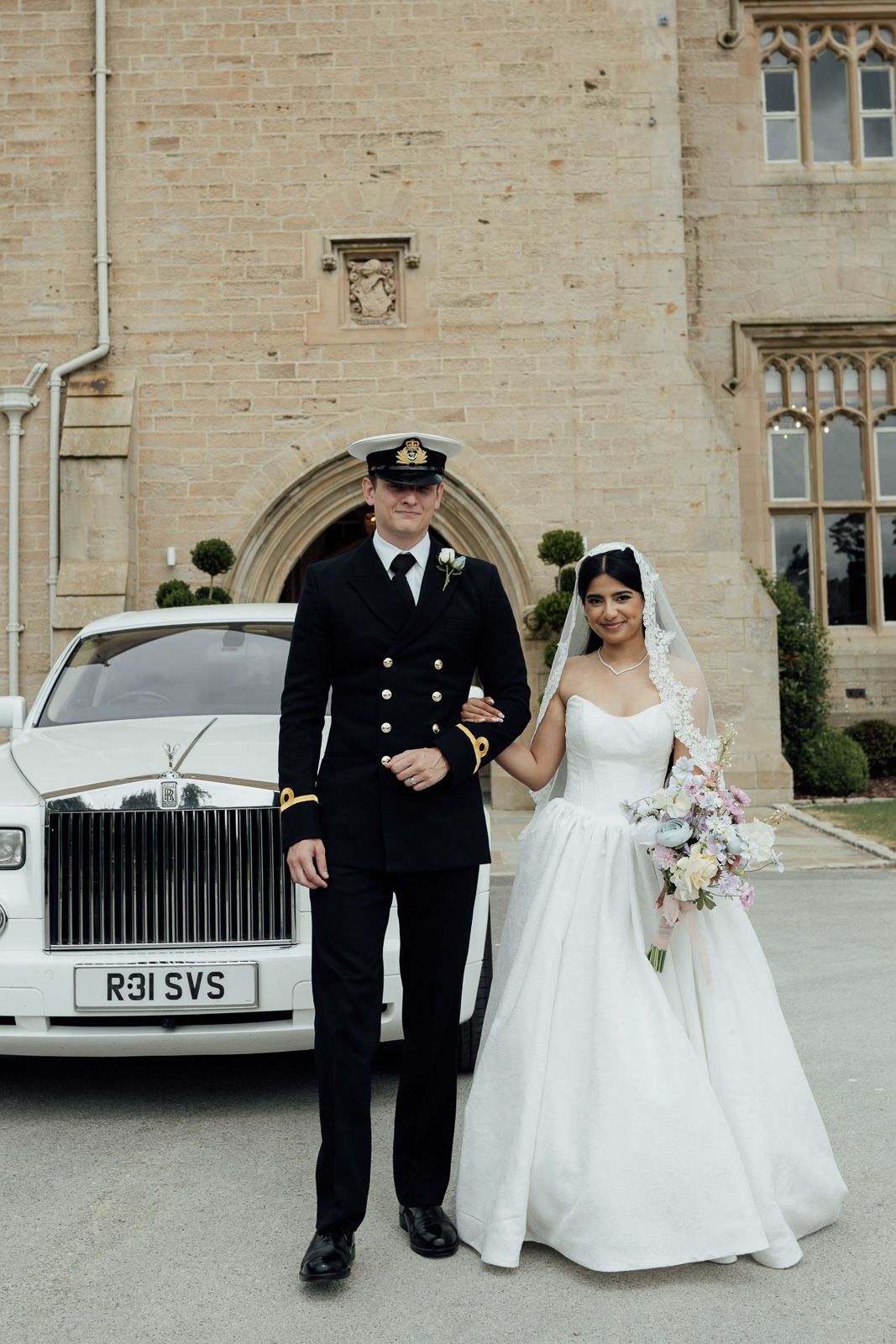 A bride and groom pose in front of a white Rolls-Royce at a castle wedding venue, he in naval uniform, she in a white gown.