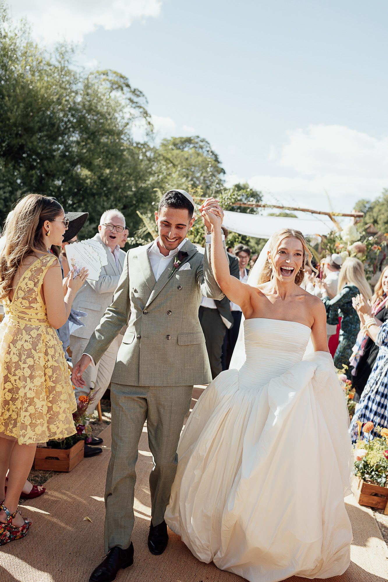 Joyful bride and groom walk down the aisle after wedding ceremony, surrounded by cheering guests outdoors.