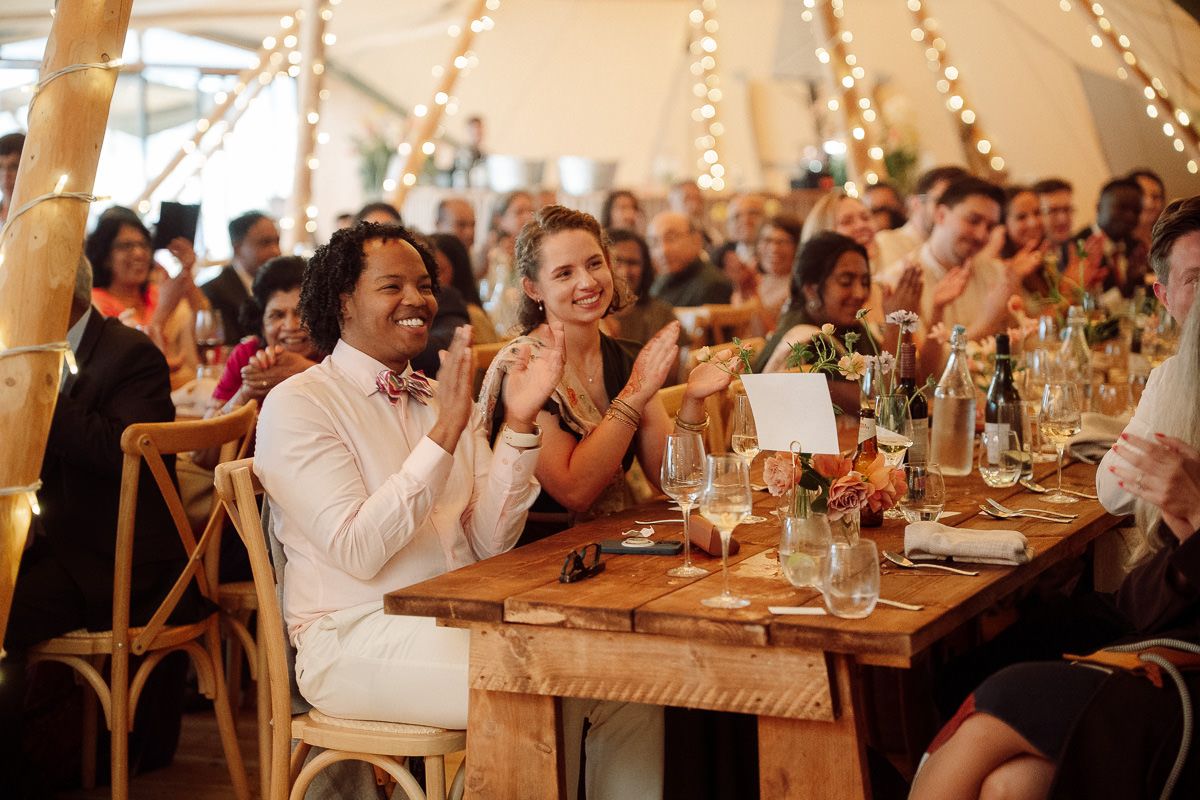 Wedding guests enjoying a celebration inside a tent with string lights and wooden tables.