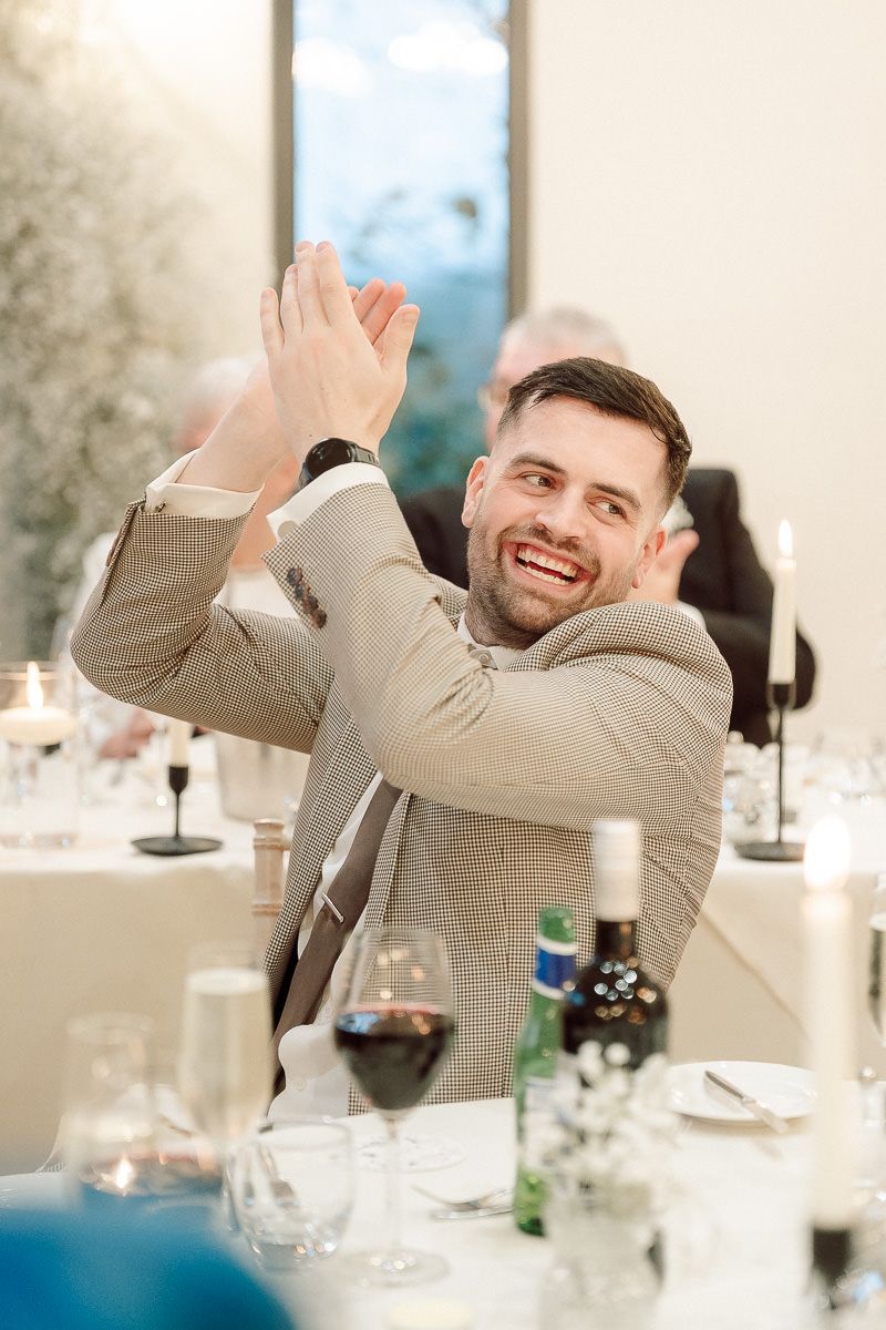 A guest in a beige suit jacket claps enthusiastically during a celebration dinner with wine glasses on the table.