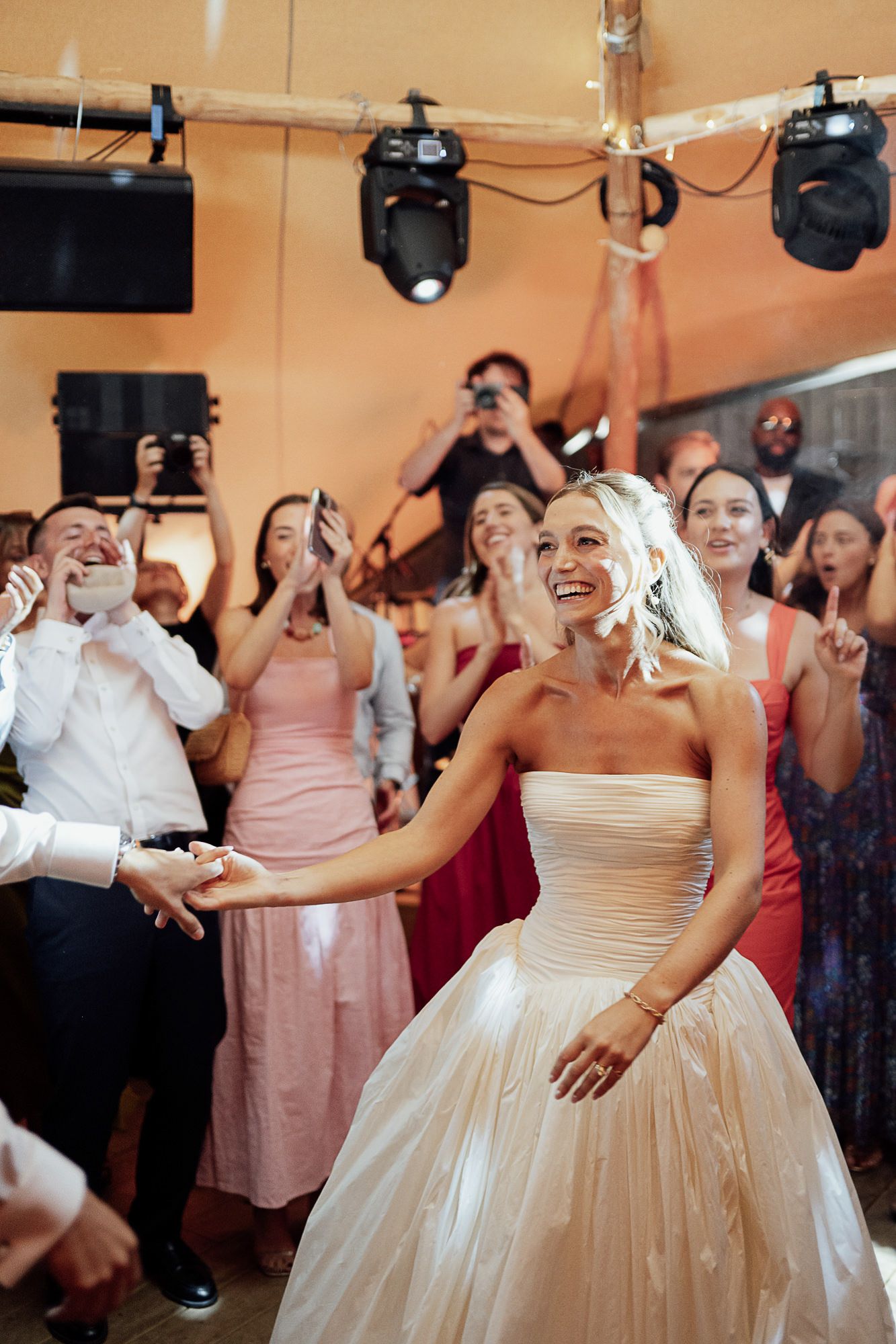 Smiling bride in strapless gown reaches out during first dance, surrounded by cheering wedding guests.
