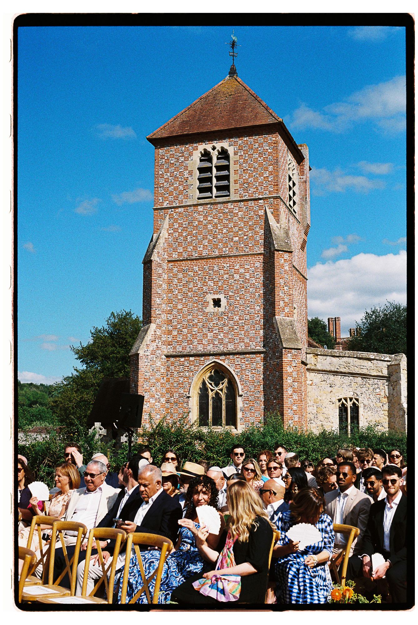 Guests seated outdoors at a sunny wedding ceremony beside a historic brick church tower with blue skies.