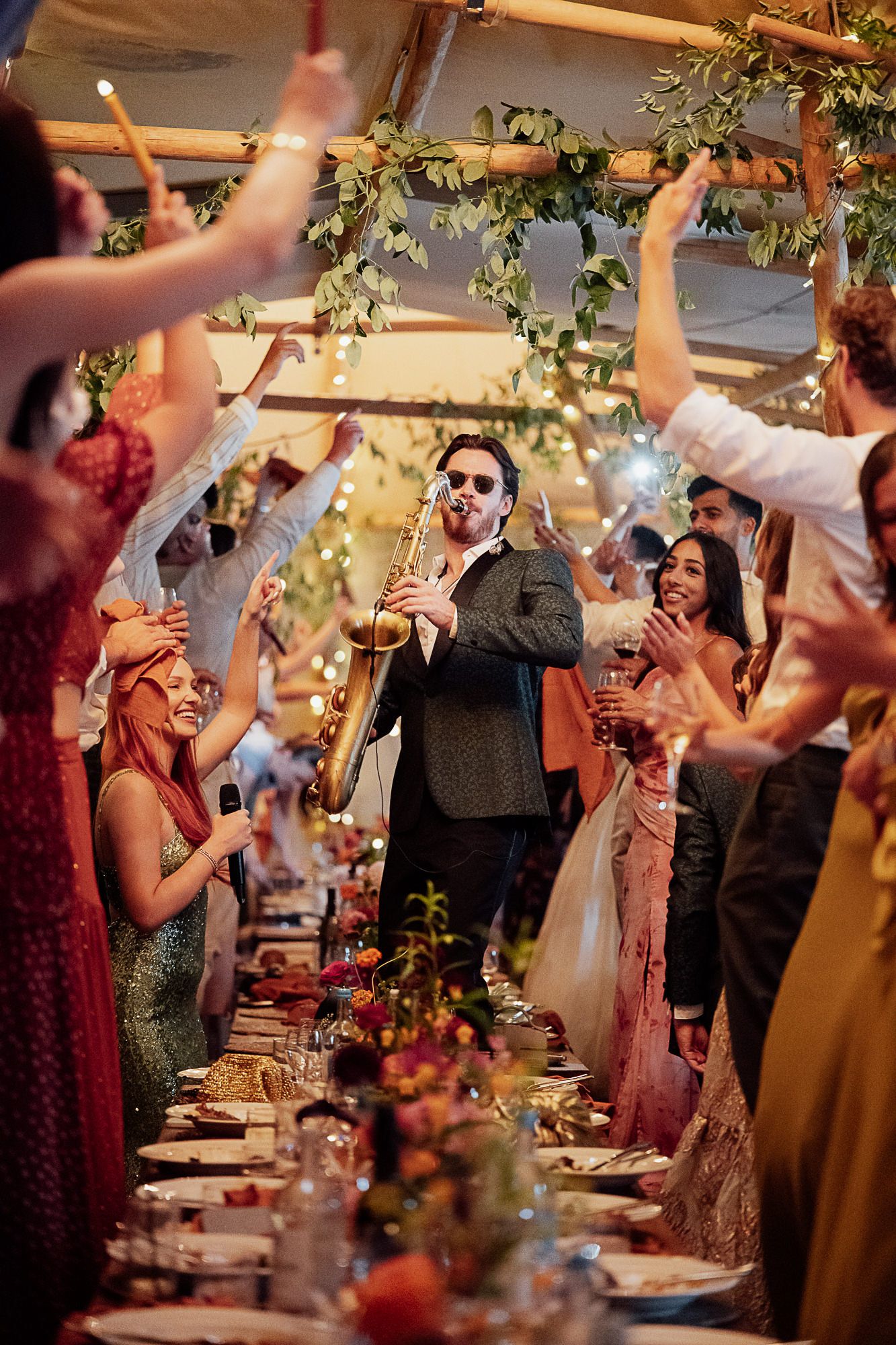 Saxophonist performing at a lively wedding reception under a decorated tent with guests celebrating around long dinner tables.