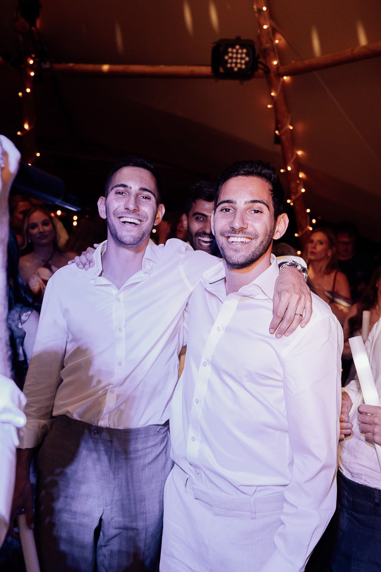 Two smiling groomsmen in white shirts celebrate together on the dance floor at a festive wedding reception.
