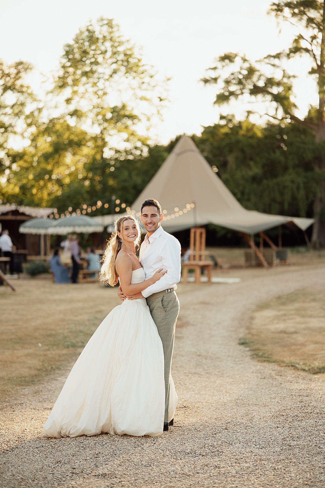 Bride and groom embrace on a sunlit path at an outdoor wedding venue with a tipi and fairy lights.