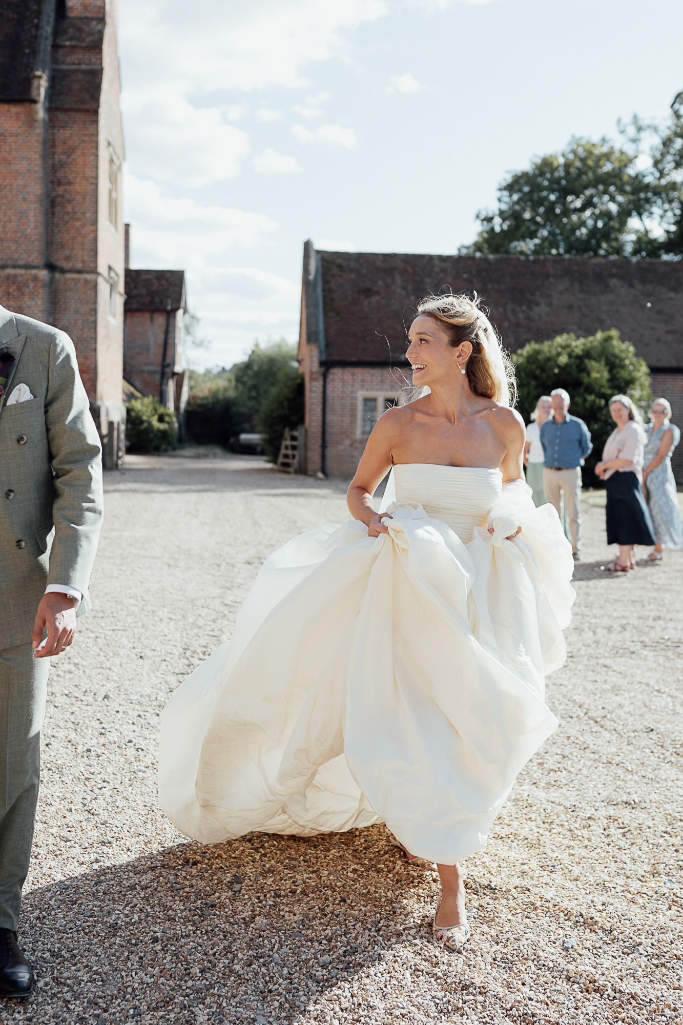 Smiling bride in a strapless ballgown walks across a gravel courtyard at a countryside wedding venue.