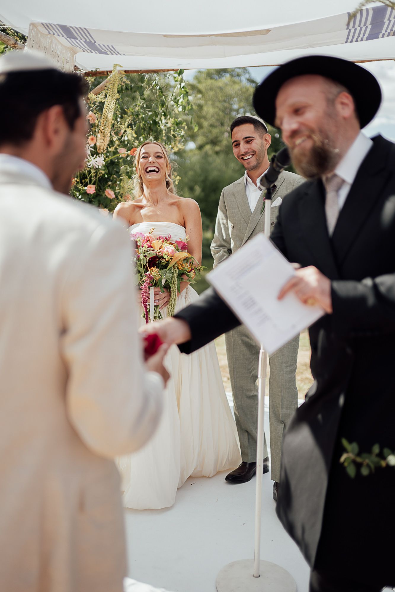 Joyful bride and groom laugh during outdoor Jewish wedding ceremony under chuppah with rabbi officiating.