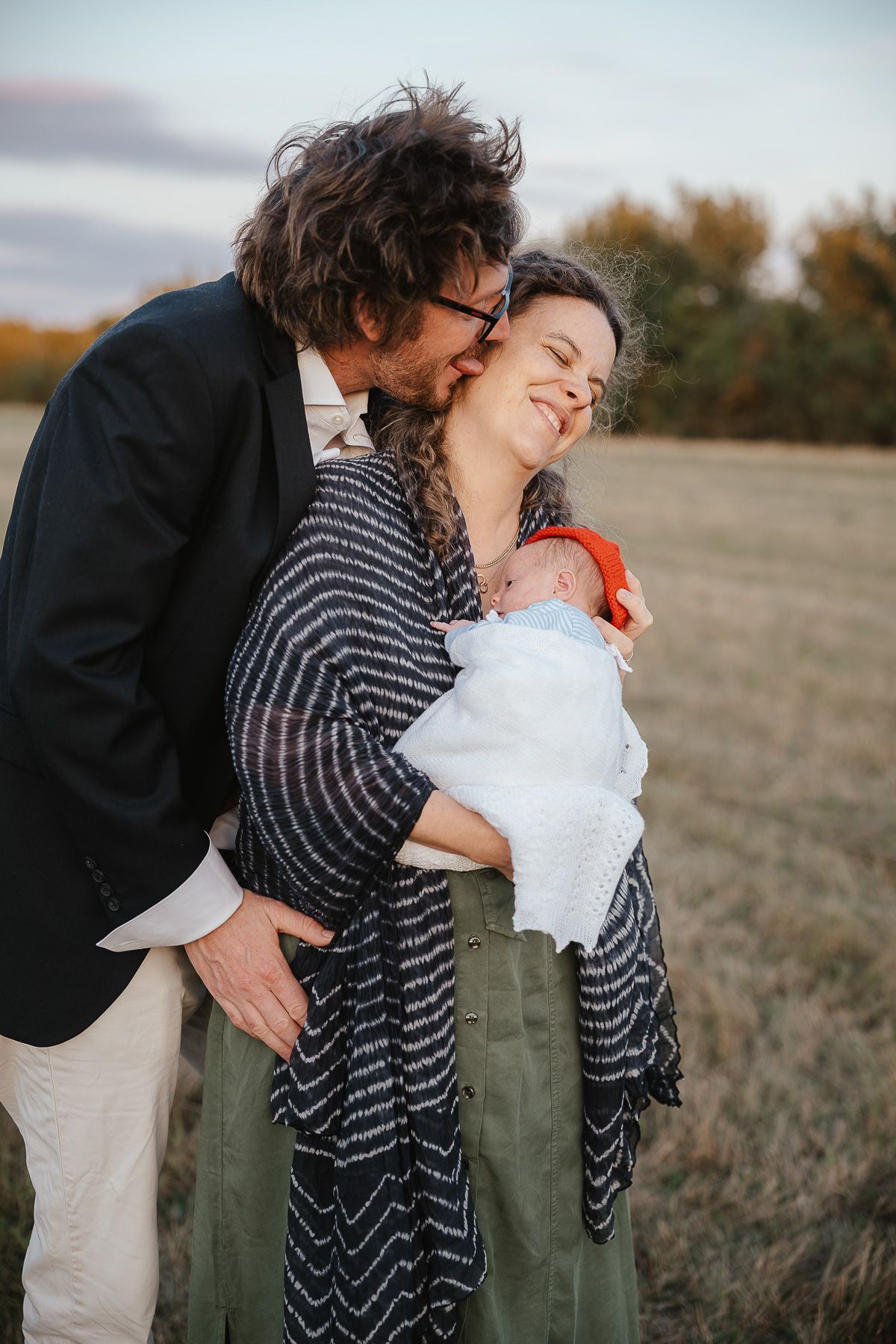 An intimate family moment captured outdoors in a field during autumn with a baby wrapped in white.