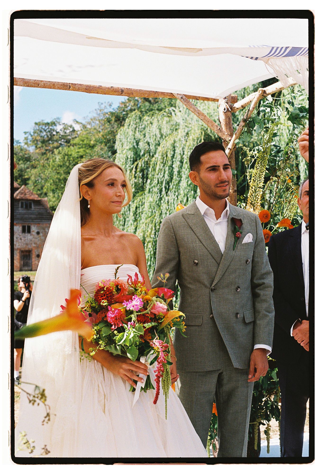 Bride in white strapless gown with colorful bouquet stands beside groom in gray suit at outdoor wedding ceremony.