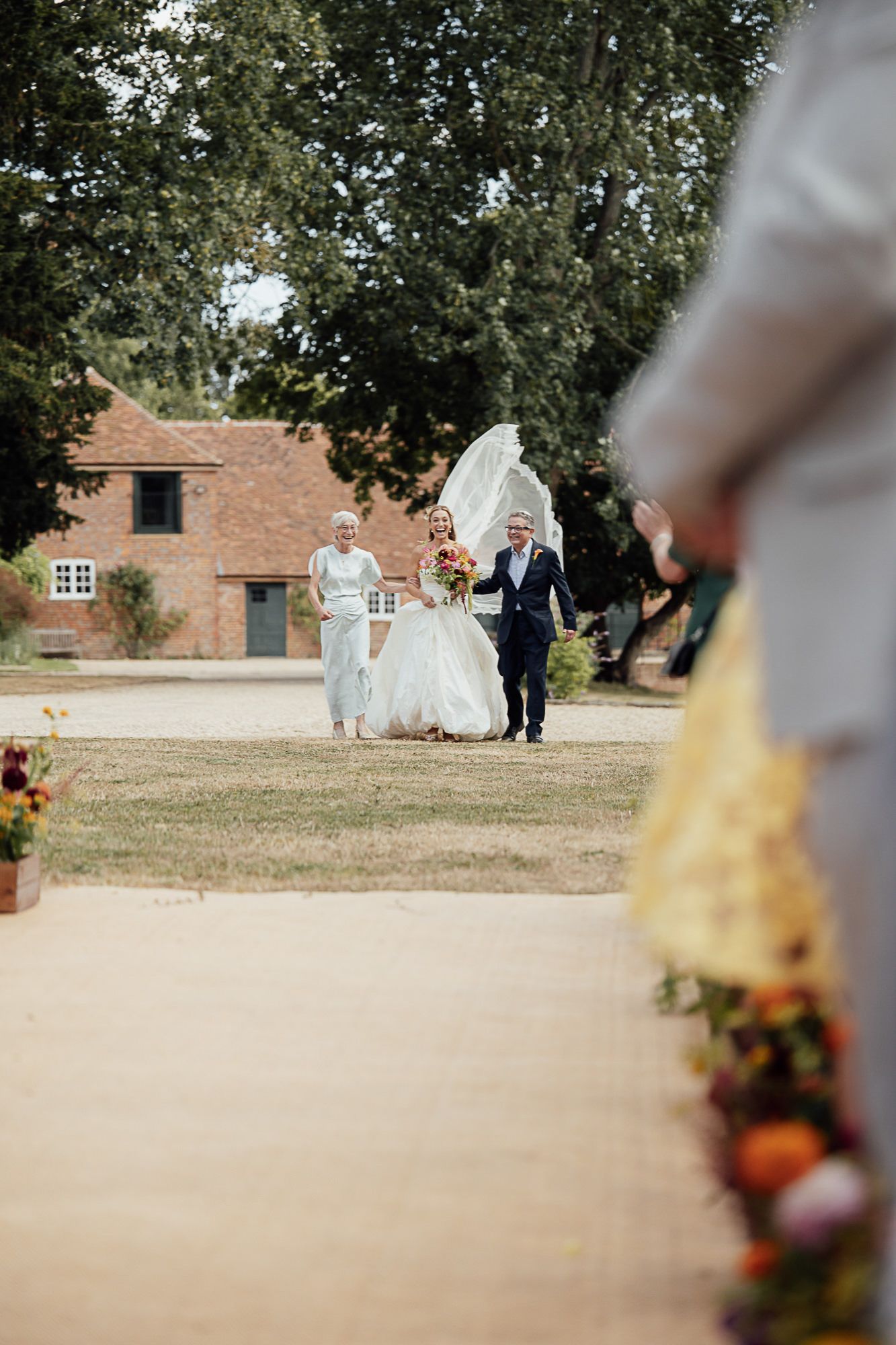Bride walks outdoor aisle with parents, veil blowing in wind, colorful bouquet, rustic barn venue in background.