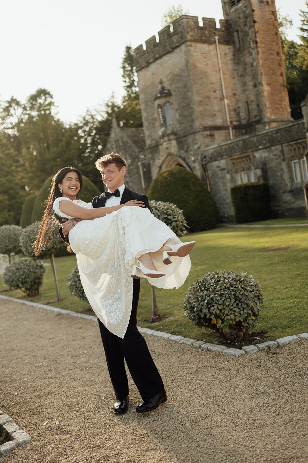 Joyful groom carries his laughing bride in a white gown outside a historic castle wedding venue.