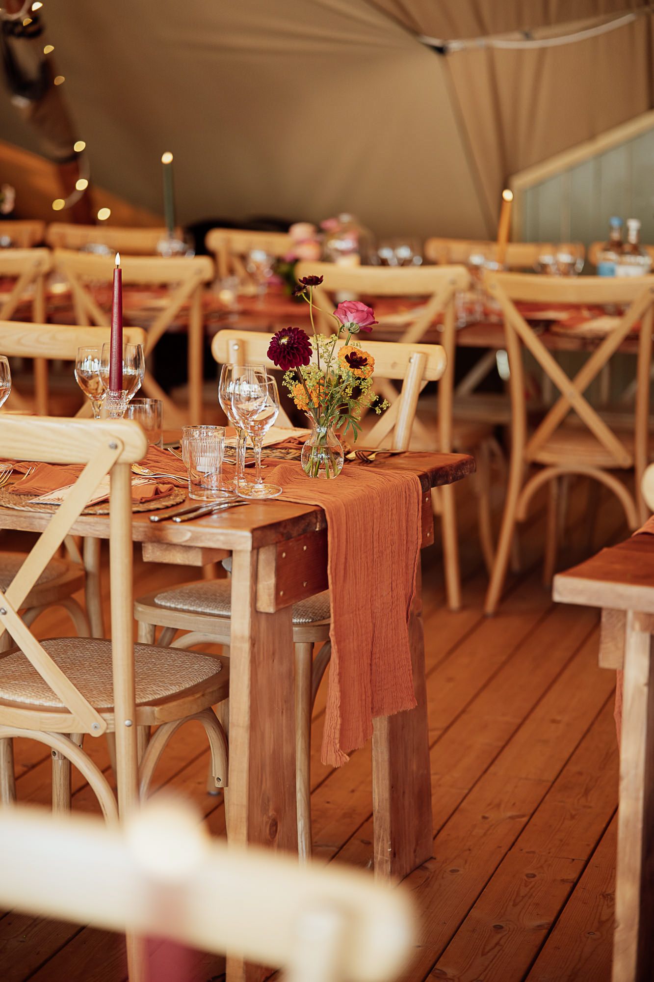 Rustic wedding reception inside a tipi tent with wooden tables, terracotta runners, floral centerpieces, and string lights.