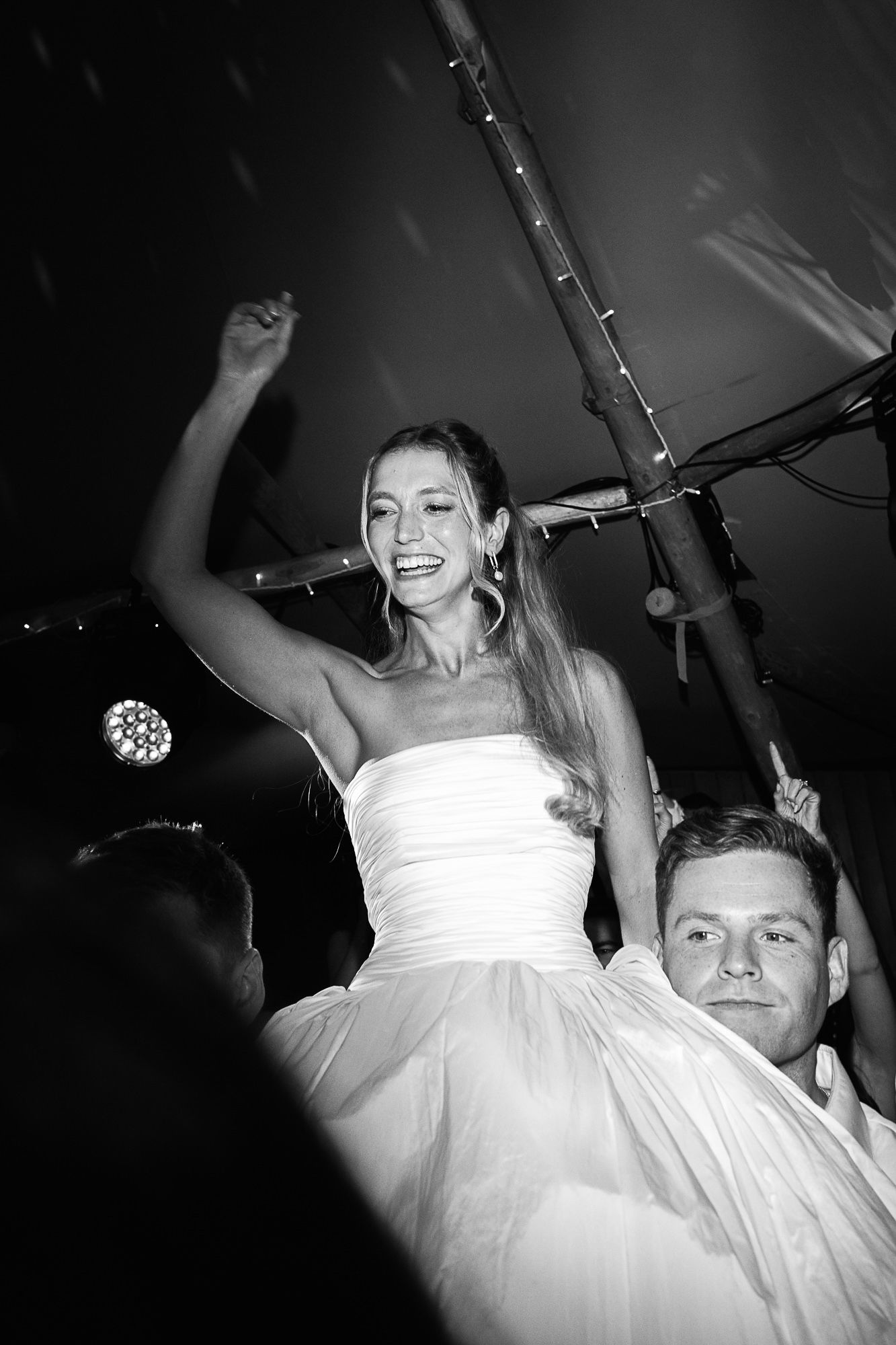 Bride in white strapless gown cheering with raised arm while being lifted by groom at wedding reception.