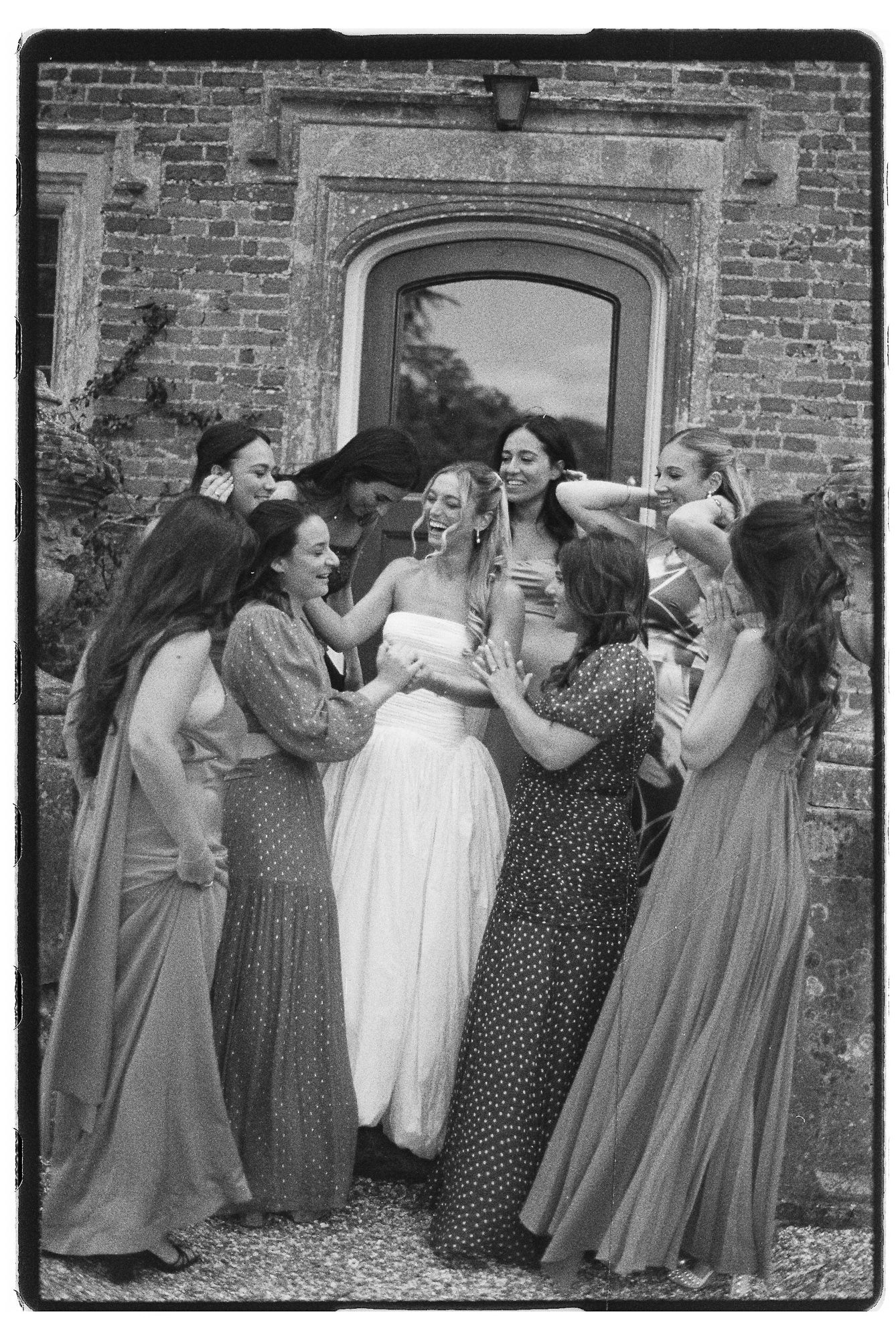 Black and white photo of bridesmaids in floral gowns laughing with bride outside brick building.